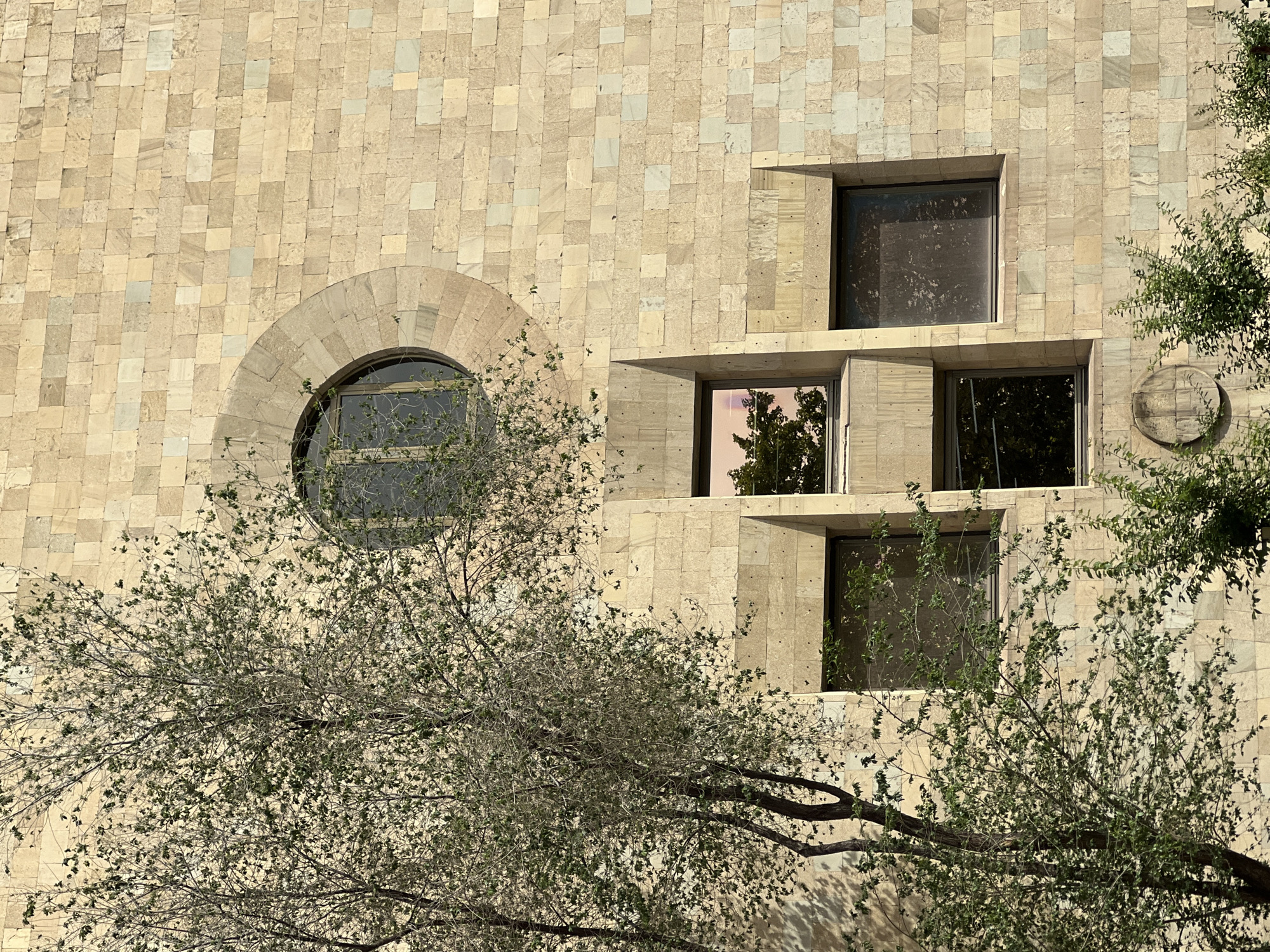 The image depicts a facade of a building constructed from light-colored, roughly square stone tiles. A round window is on the left of the building's facade. A series of square windows are stacked vertically in the center and right of the facade. Some windows reflect foliage. A tree with sparse leaves partially obscures the lower portion of the building. A circular architectural detail is visible on the right of the facade.
The description does not contain any information about the location in Yerevan, Armenia, as it is not discernible from the image.