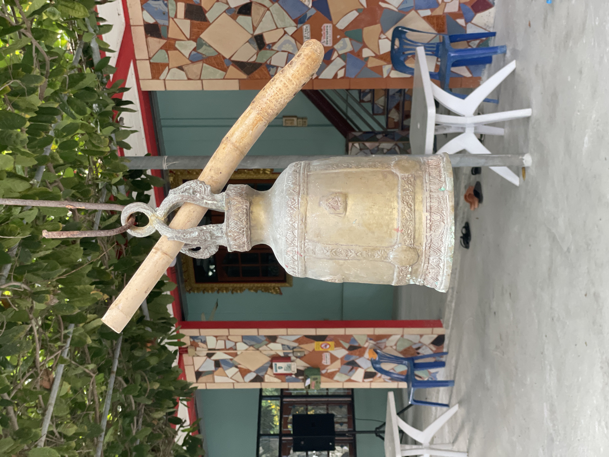 An aged, bronze bell hangs from a metal rod, suspended by a rusty hook beneath a canopy of green leaves. The bell, adorned with intricate patterns and decorative bands, rests in the center of the frame. To the left, a white table and a blue plastic chair are visible. To the right, another white table and a blue chair are situated, with a staircase and a wall covered in colorful mosaic tiles in the background. A black speaker on a stand is partially visible to the left, and a few pairs of sandals are scattered on the concrete ground. The scene appears to be an outdoor area in Pattaya City, Thailand, possibly a temple or a public space.
