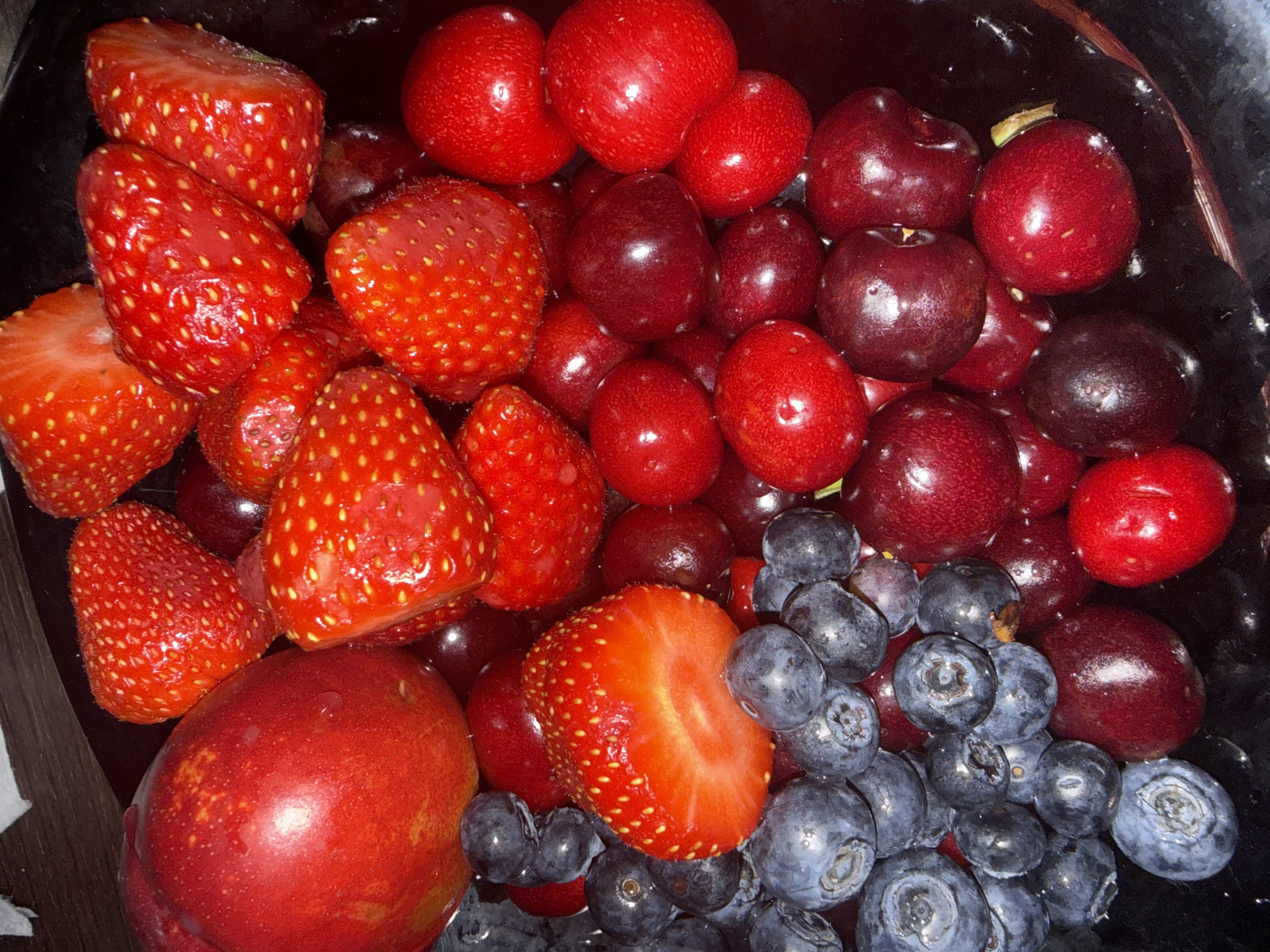 A high-angle, close-up shot shows a dark bowl filled with fresh fruit. Strawberries, cherries, blueberries, and a nectarine or plum are visible. The strawberries are bright red and some are cut in half. The cherries range in color from bright red to dark burgundy. The blueberries are clustered together, displaying a dusty blue-gray hue. A single red nectarine sits amongst the other fruit. The bowl appears to be inside, possibly a kitchen. There is no indication of the location being Sofia, Bulgaria. There are no people, subjects, actions, or interactions observed. The focus is solely on the fruit and the bowl containing it.