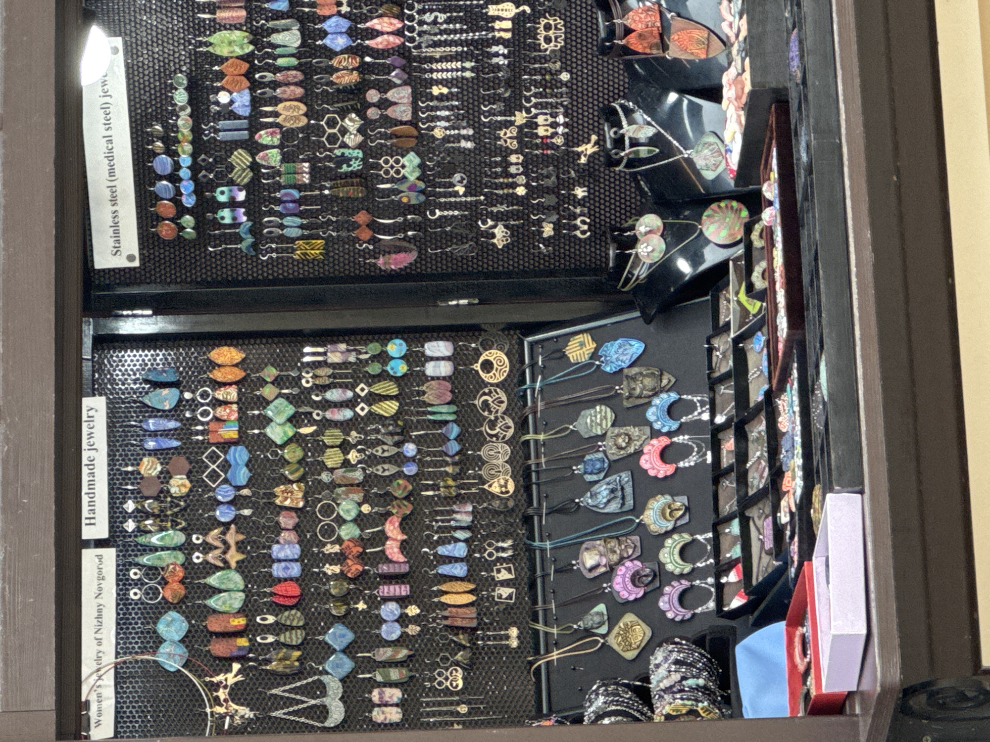 The image shows a display of handmade jewelry in a shop located in Nizhny Novgorod, Russia. The display features a variety of earrings and necklaces, with a focus on unique, handcrafted designs. The jewelry is arranged on a black mesh background, with some pieces hanging and others displayed on mannequins. The shop appears to be a small, independent boutique, likely catering to tourists or locals looking for artisanal goods. The lighting is somewhat dim, but the jewelry is clearly visible. There are no people visible in the image.