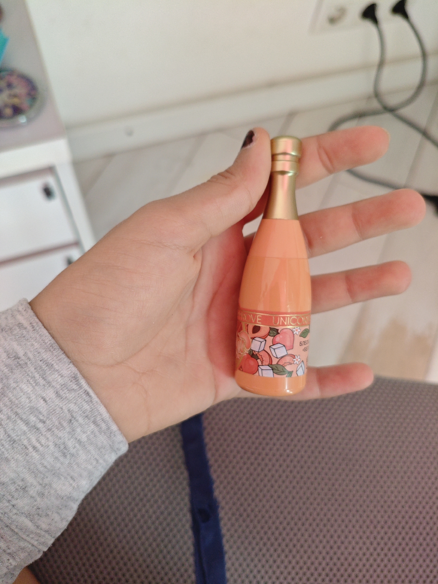 A hand holds a small, orange, bottle-shaped object with a gold cap. The bottle has a label with an illustration of peaches, ice cubes, and leaves. Text on the label is partially visible, reading "ROVE UNICORN" and some Cyrillic text. The background is out of focus, showing a white wall with electrical outlets and cords, and a glimpse of a piece of furniture with a patterned surface. The image appears to be taken indoors. There is no indication of the location being in Трудовское сельское поселение, Ukraine.
