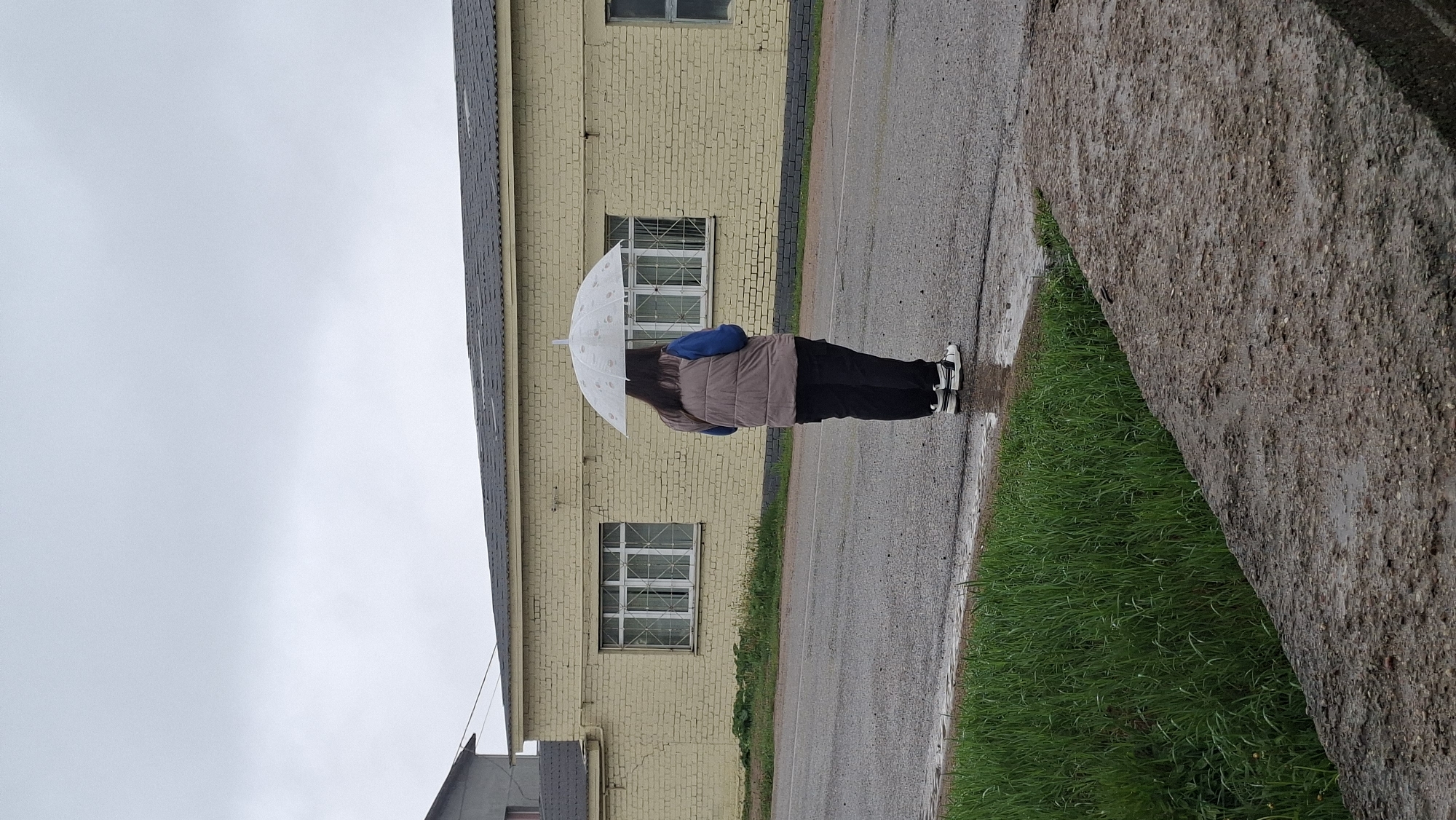 The image depicts an individual standing under a white umbrella on a wet asphalt road in Unknown, Unknown. The person is wearing a taupe vest, a blue garment underneath, and black pants. They are also wearing white shoes with black accents. Adjacent to the road is a grassy area, bordered by a concrete structure. A yellow brick building with white-framed windows is in the background. The sky is overcast.