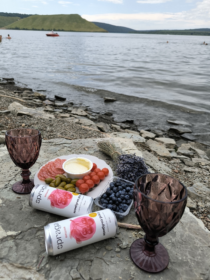 The image displays a detailed picnic scene situated on a rocky shoreline in Horaivka, Ukraine. In the immediate foreground, a cluster of items is arranged on flat, greyish-brown rock formations. These items include two dark purple, patterned glass goblets positioned on the left and right sides. Centrally, a white scalloped plate holds an assortment of food: slices of reddish sausage or salami, small green pickles, and a pile of red cherry tomatoes. A small white bowl on the plate contains a yellowish, creamy substance. Directly in front of the plate are two white aluminum cans, identified as "spraga" kombucha with cherry illustrations. To the right of the plate, a clear plastic container is filled with blueberries. Beside the blueberries, a small, tied bundle of dried purple lavender flowers is visible, alongside a small bundle of dried straw or grass.
The midground transitions to the water's edge, where small waves lap against the stony shore, which is composed of numerous flat, grey rocks. The background features a large body of water, consistent with the Dniester River, extending into the distance under a partly cloudy sky. Several individuals are observed swimming or wading in the water. A red motorboat with a visible wake is traversing the water from left to right in the mid-distance. Across the water, distant green, rolling hills are prominently visible.
