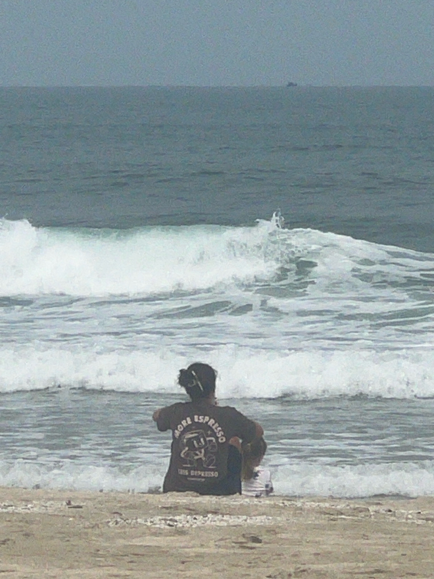 The image shows a person sitting on a beach facing the ocean. The person is wearing a dark t-shirt with a graphic design and text. The ocean has moderate waves and the sky is overcast. There are no discernible city landmarks or specific features of Đà Nẵng visible in the image.