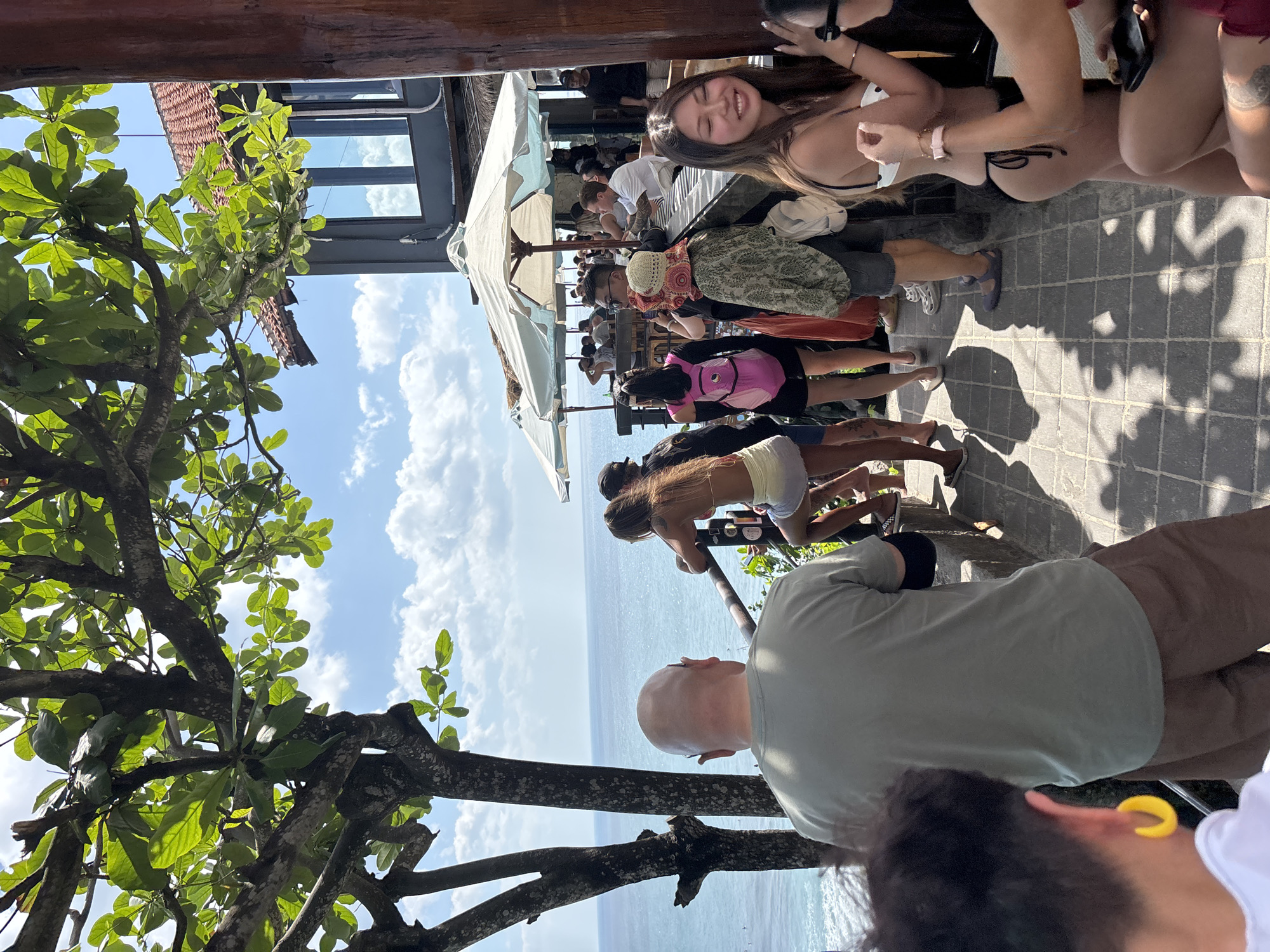 The image captures a bustling cliffside scene overlooking the ocean, likely in Kuta Selatan, Indonesia, during bright daylight hours. The foreground is dominated by the dense green foliage and thick branches of a large tree on the left, which partially frames a clear blue sky with scattered white cumulus clouds.
In the immediate foreground, from the left, a person with short dark hair and a prominent yellow hoop earring is partially visible from behind. Slightly to their right and central, a bald man wearing a light olive-green t-shirt and brown shorts stands facing the ocean, his back to the viewer. He has a dark sleeve or band on his left arm.
To the right of the bald man, along a dark metal railing that runs parallel to the cliff edge, several individuals are visible. One woman with long, light brown hair, wearing a light-colored bikini top and light shorts, leans on the railing, looking out towards the sea. Further right, a young woman with long dark hair, wearing a white polka-dot bikini top and black bikini bottoms, smiles broadly, looking towards the right of the frame. She wears a light pink watch or bracelet on her left wrist and holds a dark smartphone.
In the mid-ground, behind the railing, a paved outdoor area hosts numerous people gathered around a bar setup. Two large white patio umbrellas provide shade over this area. Many individuals are either standing or seated on high stools at the bar counter, engaging in conversation or looking out at the ocean. Notable figures include a man in a white t-shirt seated at the bar and a woman with a pink backpack facing the ocean. Distinct shadows of people and tree leaves are cast across the grey patterned pavement, indicating strong sunlight.
In the background, to the right, a multi-story building features a reddish-brown tiled roof, dark grey walls, and large reflective windows that mirror the sky and clouds. The vast blue ocean, with visible white surf near the distant shore, stretches to the horizon under the bright sky. The overall scene depicts a lively, social outdoor setting with panoramic views.