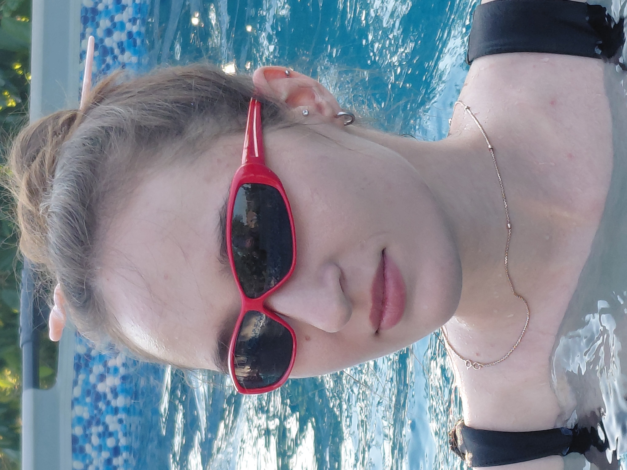 A young person with light brown hair pulled back is partially submerged in a swimming pool. They are wearing red sunglasses with dark lenses and a thin gold chain necklace. Their ears are pierced with multiple small rings, and a pink hair clip is visible in their hair. The water is a bright blue and appears clear, with ripples on the surface reflecting light. The background of the pool is a mosaic of blue and white tiles. The person's skin is damp, suggesting they have been in the water for some time. They are wearing a dark-colored top, possibly a swimsuit, with black bands visible around their upper arms. The image is taken from a close-up perspective, focusing on the person's face and upper body. The lighting suggests it is daytime, and the overall mood is relaxed and summery.