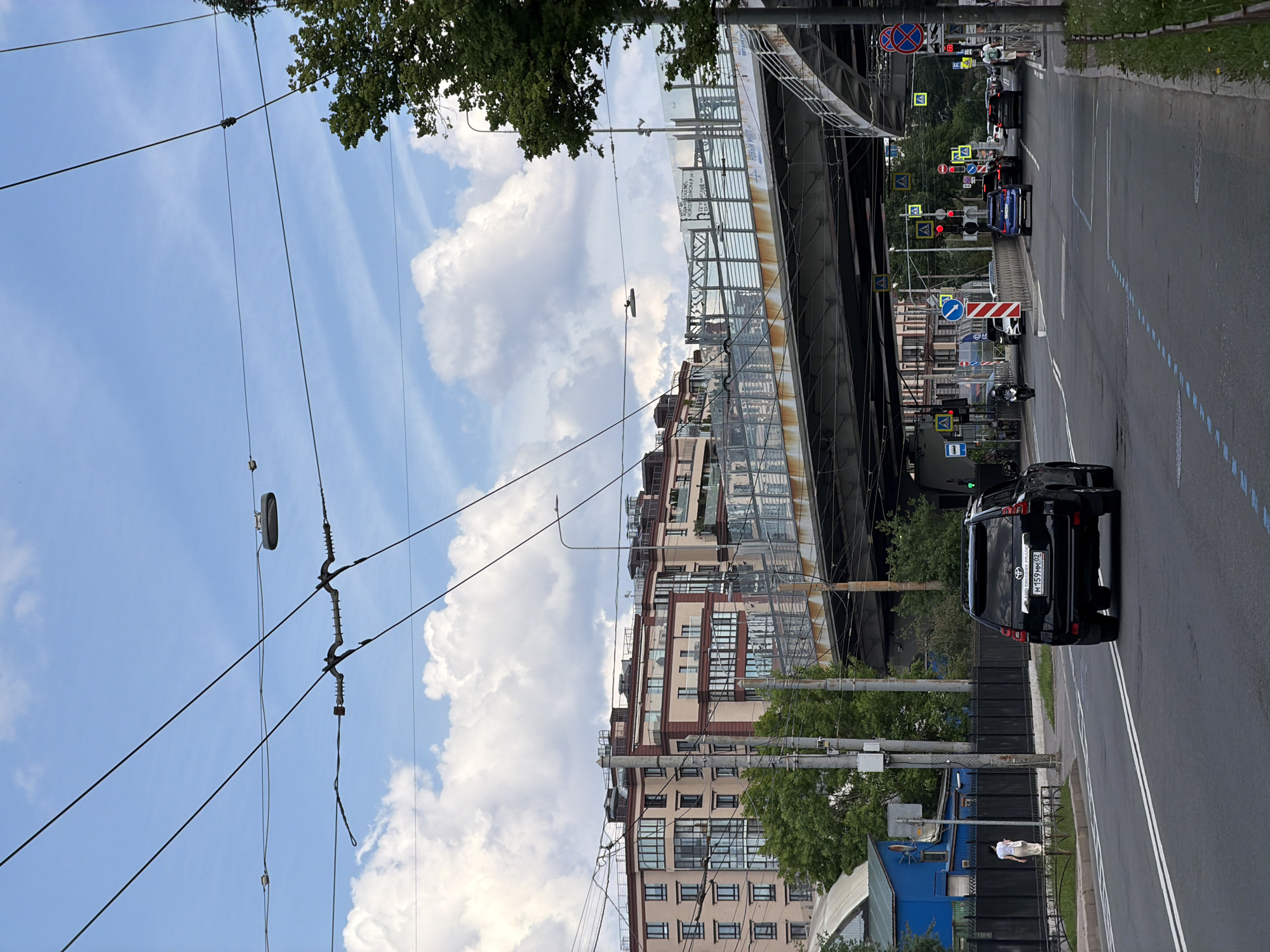 This image depicts an urban street scene at the intersection of Novoizmailovsky Prospekt and Blagodatnaya Street in Saint Petersburg, Russia, specifically showing the underside and approach to the Blagodatny Bridge.
In the foreground, an asphalt multi-lane road features white solid and dashed lane markings, with additional blue dashed lines in the left lane. A black Toyota Land Cruiser Prado SUV, bearing Russian license plate M159MM02, is positioned in the rightmost visible lane, facing away from the viewer.
To the left of the SUV, a paved sidewalk abuts a black metal fence. A person in a light top and skirt walks on this sidewalk, moving away from the camera. Behind the fence, a utility pole stands near a low blue-painted building with a dark roof.
To the right of the SUV, another sidewalk borders a grassy median and a lamppost. Further down the street, multiple vehicles including a blue sedan, a dark sedan, and a white car are present at an intersection, with traffic lights displaying red. A motorcycle is visible to the far left of the intersection. A person with a backpack and what appears to be a kick scooter is walking on the right-hand sidewalk. Numerous standard Russian road signs are visible, including pedestrian crossing signs (yellow diamond with walking figure), a "no stopping" sign (red circle with a blue cross), and a bus stop sign. A leafy green tree occupies the far right side of the frame.
Dominating the midground is the Blagodatny Bridge, a large concrete and metal overpass spanning the roadway. Its underside is dark gray, with visible rust on lower metal support structures. The bridge's upper barrier consists of a metal framework with translucent panels, to which banners are attached. Partial Cyrillic text "НОВЫЙ МИР" (New World) and "НОВОГОДНЯЯ И H" (New Year and H) is discernible on one banner.
In the background, beyond and to the left of the bridge, are modern multi-story residential buildings, part of the "Moskovsky Kvartal" complex. These structures feature light-colored (pinkish-beige) facades, dark-framed windows, multiple balconies, and some reddish-brown architectural accents.
Numerous utility poles and a dense network of black overhead wires, indicative of a trolleybus or tram system, crisscross the light blue sky which is dotted with scattered white cumulus clouds. Multiple streetlights are mounted on poles and on the bridge structure. The overall scene is illuminated by daylight.