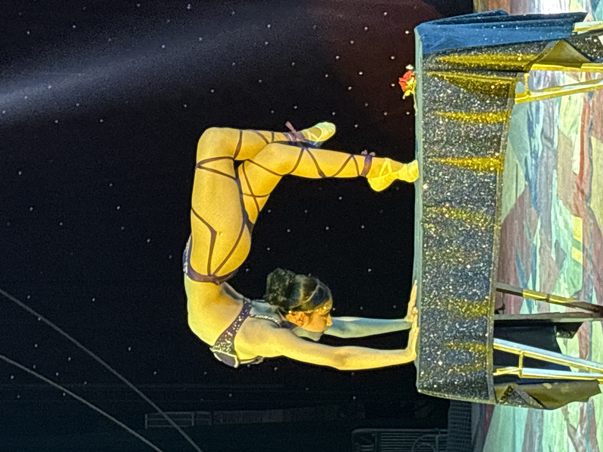 An acrobat performs on stage. The performer, a woman, is suspended from a rectangular apparatus covered in glitter. She wears a light-colored costume with purple accents and matching wrappings on her legs and feet. Her body is contorted in a bent position, using her arms to support herself. The background is dark with small white dots. The apparatus is next to a colorful painted wall. The location is Tejalpa, Mexico.