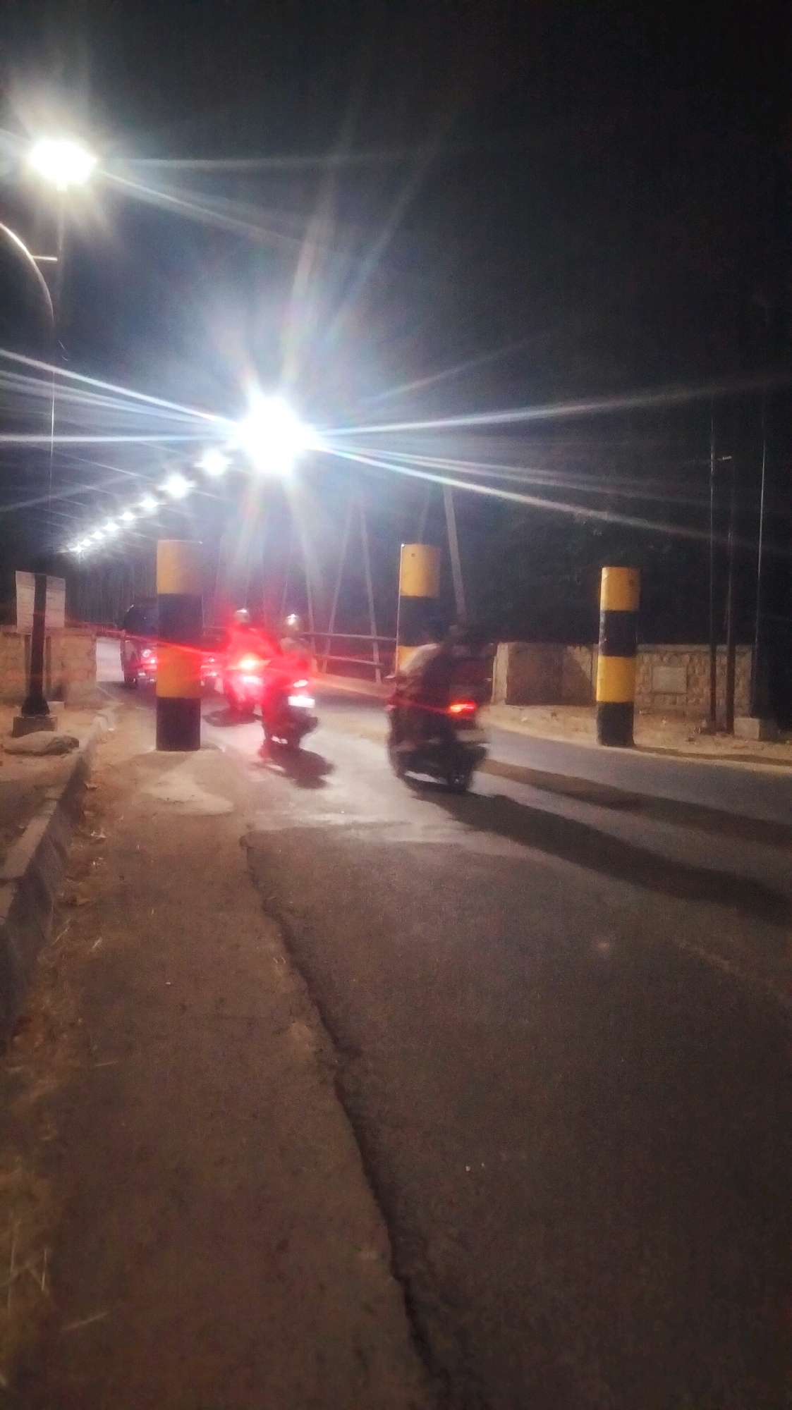 The image depicts a night scene on a road, possibly a toll booth or checkpoint, in Bojonegoro, Indonesia. Several motorcycles are visible, some with riders, on a paved road. The motorcycles are in motion, creating a blur effect, and their headlights and taillights are illuminated.
Two large, cylindrical bollards with alternating yellow and black bands are prominent on either side of the road, serving as markers or barriers. In the background, a structure resembling a bridge or overpass is visible, lined with lights that create a starburst effect due to the night exposure. The overall lighting is dominated by bright artificial lights, likely streetlights, which cast long shadows on the road surface.
The time of day is clearly night, and the weather appears to be clear. The presence of motorcycles and the setting suggest a transportation hub or a point of entry/exit. There is no visible text in the image. The atmosphere is dynamic due to the moving vehicles and the stark contrast between light and shadow.