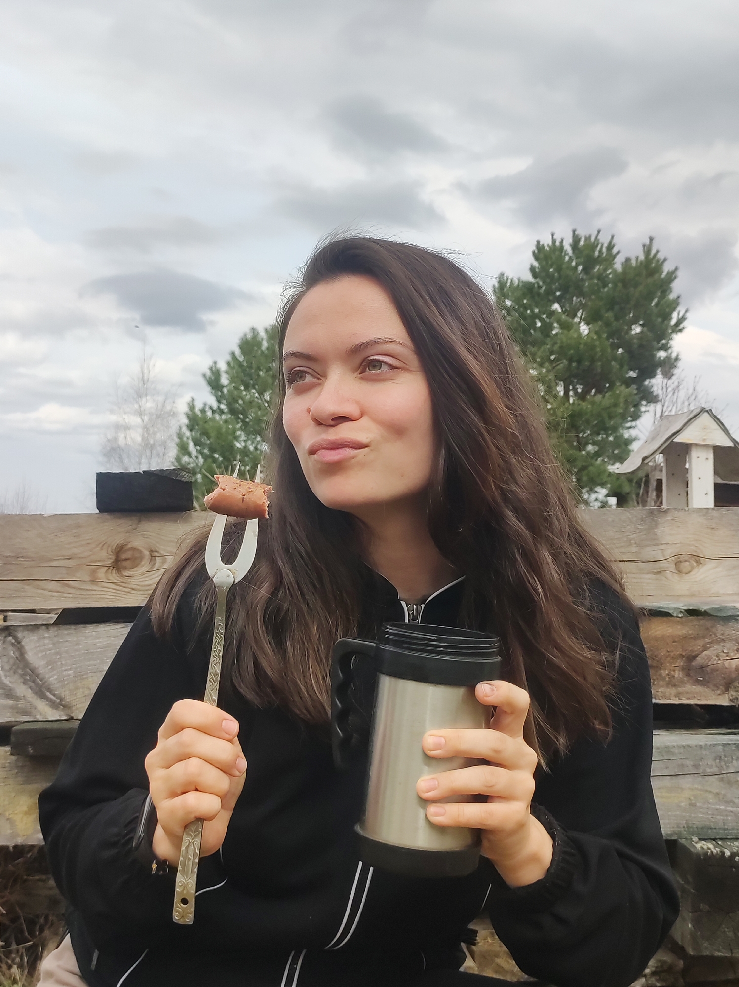 A young woman is sitting outside, holding a fork with a piece of cooked sausage and a metal thermos. She is wearing a black jacket with white stripes. Behind her is a wooden fence, trees, and a building. The sky is cloudy. The location is most likely not in the city of Фоки, Russia, as the image does not contain any identifiable features specific to that location.