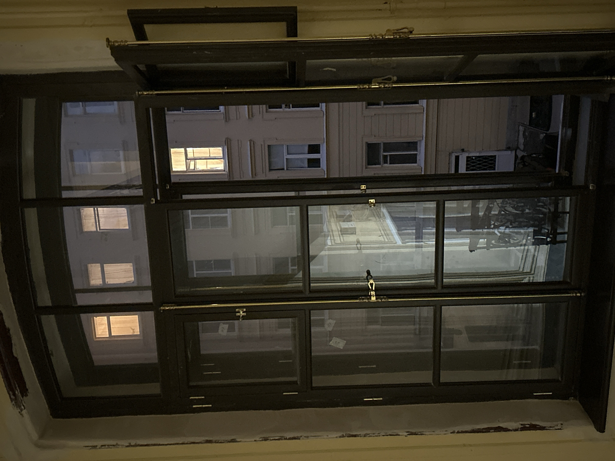 A night view through a multi-paned window shows an adjacent building with lit windows. The window is framed by dark wood, with brass hardware securing the open sections. In the foreground, the window panes reflect the interior of the room. The adjacent building is several stories high, with architectural details like cornices and pilasters. Some windows have curtains, while others appear empty. The overall lighting suggests dusk or night, with artificial light emanating from within the buildings.
