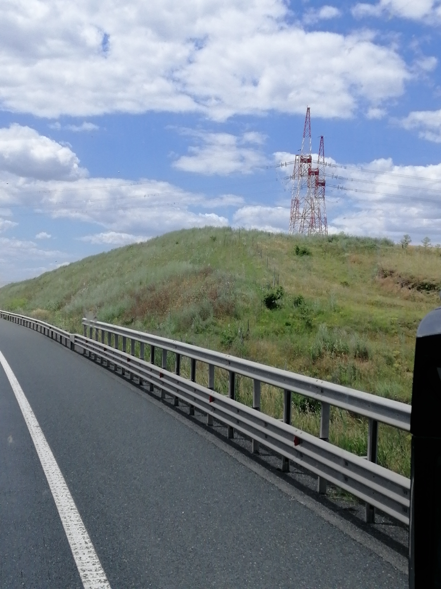 The image depicts a highway scene in Făclia, Romania. The foreground features a section of an asphalt road with a white lane marker. To the right of the road is a metal guardrail. Beyond the guardrail, a grassy, undulating hill rises, dotted with sparse vegetation and a few small trees.
Dominating the background is a tall, red and white lattice transmission tower, a significant piece of infrastructure. The tower is topped with multiple crossbars supporting power lines that extend out of the frame. The sky above is blue with scattered, fluffy white clouds, suggesting fair weather during daytime. The overall impression is of a rural or semi-rural setting, likely during the day. No people are visible in the image, and no text is present. The scene conveys a sense of travel or passage through a landscape characterized by open spaces and utility infrastructure.