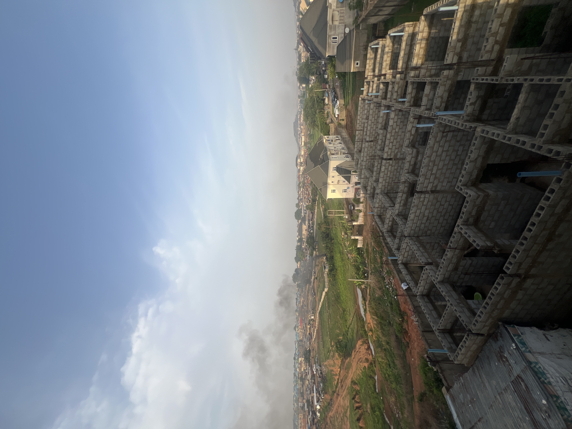 The image shows a cityscape taken from an elevated position, possibly a high-rise building under construction. The sky is visible with a mix of clear blue and hazy, cloudy areas. A significant portion of the cityscape is covered in what appears to be smoke, possibly indicating a fire or burning. The surrounding environment consists of residential buildings of various sizes and conditions, ranging from simple structures to more modern and elaborate designs. Some green vegetation is present in the open areas.
The foreground features the unfinished construction of a multistory building, characterized by exposed brickwork and steel reinforcing. Blue plastic pipes are visible within the structure.
The location is identified as Abuja, Nigeria, based on common architectural styles and environmental characteristics often seen in that region. No people are directly visible in the image.