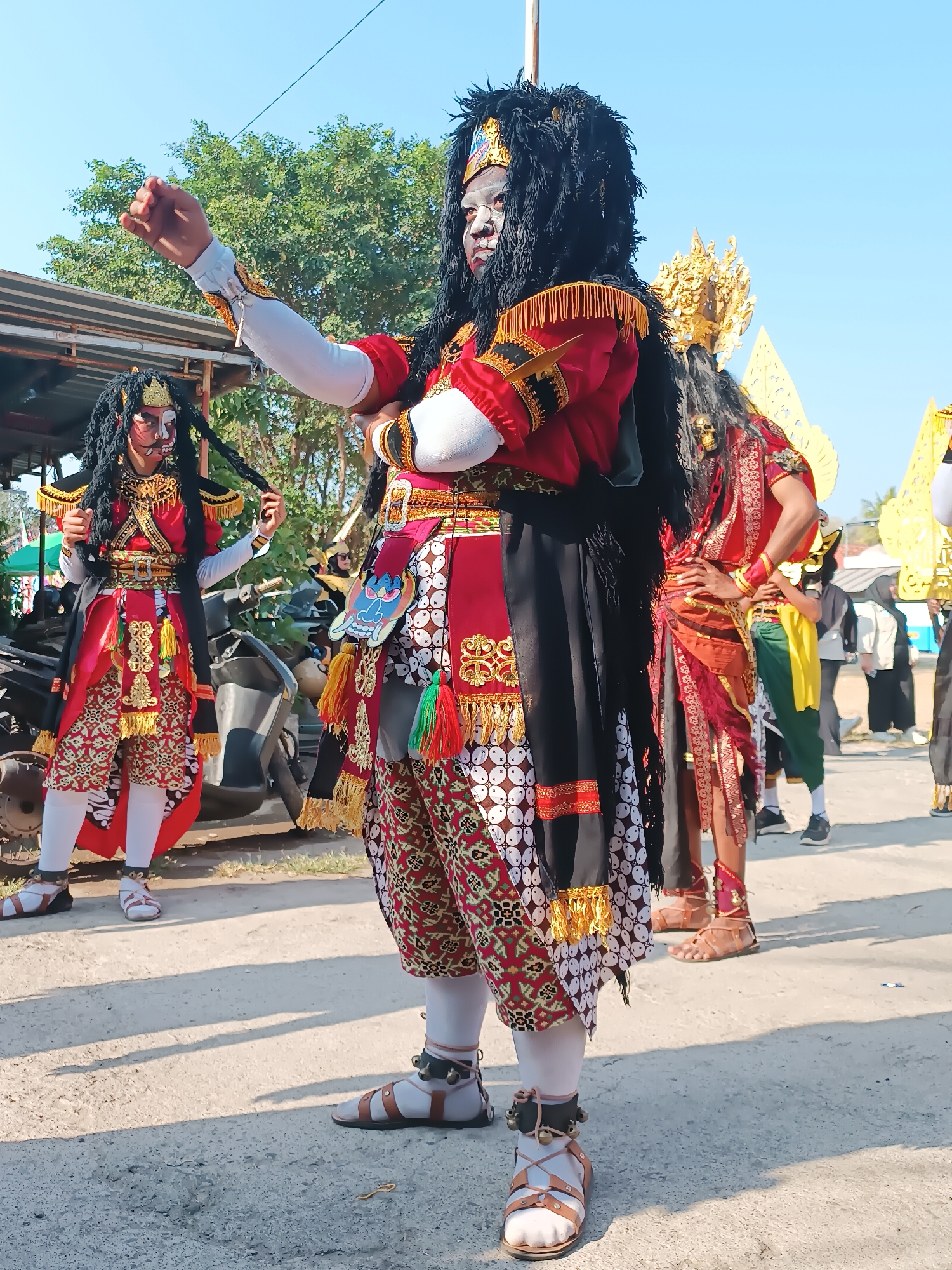 In Imogiri, Indonesia, two individuals in elaborate traditional costumes stand in what appears to be a street or open area. The person in the foreground, on the right, is the most prominent. They wear a black shaggy wig and a face mask painted white with red accents around the eyes and mouth. Their costume is a vibrant combination of red, black, and gold, featuring intricate patterns and gold trim. A large black fabric hangs from their shoulders, reaching down their back. They have their arms crossed, with their right arm extended slightly forward. Their legs are clad in white leggings and adorned with detailed sandals that have straps and bells.
To the left, a second individual, also in a similar elaborate costume with a mask, is slightly behind and to the left of the first. This person has long black braided hair extending from their mask and is holding one of the braids. Their costume is also red and gold with detailed patterns.
In the background, several other people are visible, some also in traditional attire and headdresses. A motorcycle is parked to the left. The environment appears to be outdoors, with a clear blue sky above and trees and buildings visible in the distance. The ground is paved with a light-colored material, possibly concrete or asphalt, with shadows cast by the figures. The overall scene suggests a cultural event or performance.