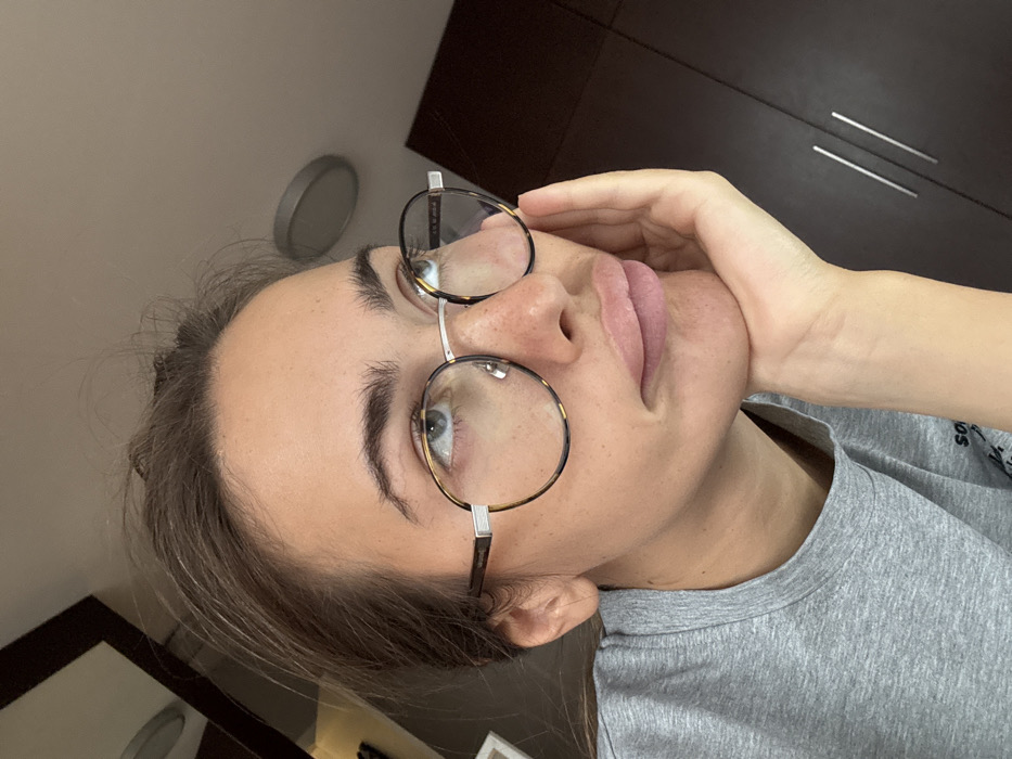 The scene is an indoor setting within the city of Warsaw, Poland. An adult female individual, with light skin and light brown hair pulled back, is depicted in a medium close-up, facing slightly right and looking upwards to the left. She wears round eyeglasses with tortoise-shell patterned frames and silver metal components. Her lips are visibly full, and light freckling is present across her nose. She is dressed in a grey crew-neck t-shirt. Her right hand supports her chin. The background features a plain light-colored ceiling with a circular recessed light fixture. Dark brown cabinet doors with two silver vertical bar handles are visible to the right, and a partial white rectangular object is visible on the far left.