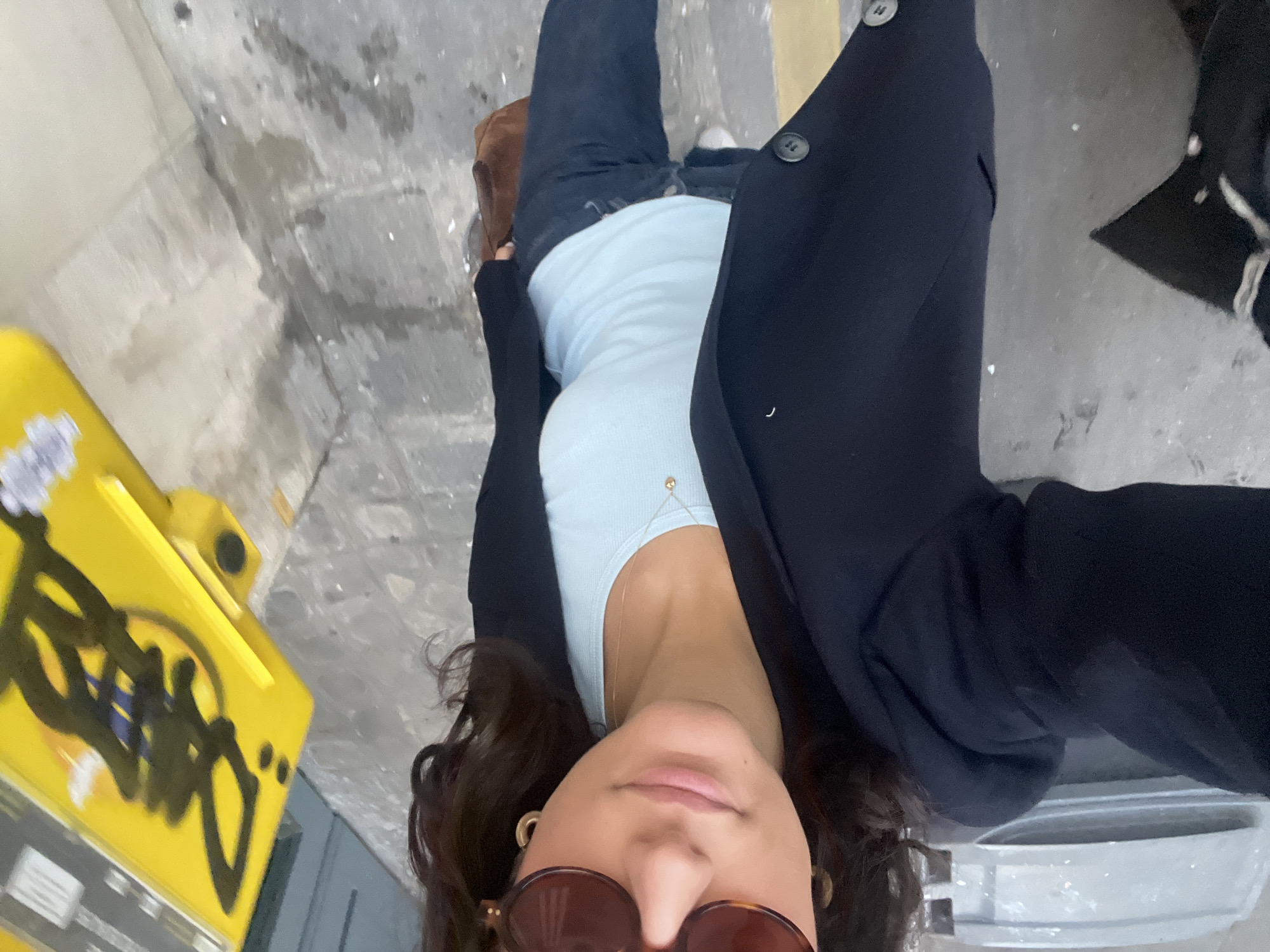 A young woman is taking a selfie outdoors. She is wearing sunglasses, a light blue tank top, a dark blazer, and dark jeans. She stands on a sidewalk next to a yellow object covered in graffiti. The setting appears to be an urban environment. Based on the style of the signs and the overall environment, it is highly probable that the location is in Paris, France.