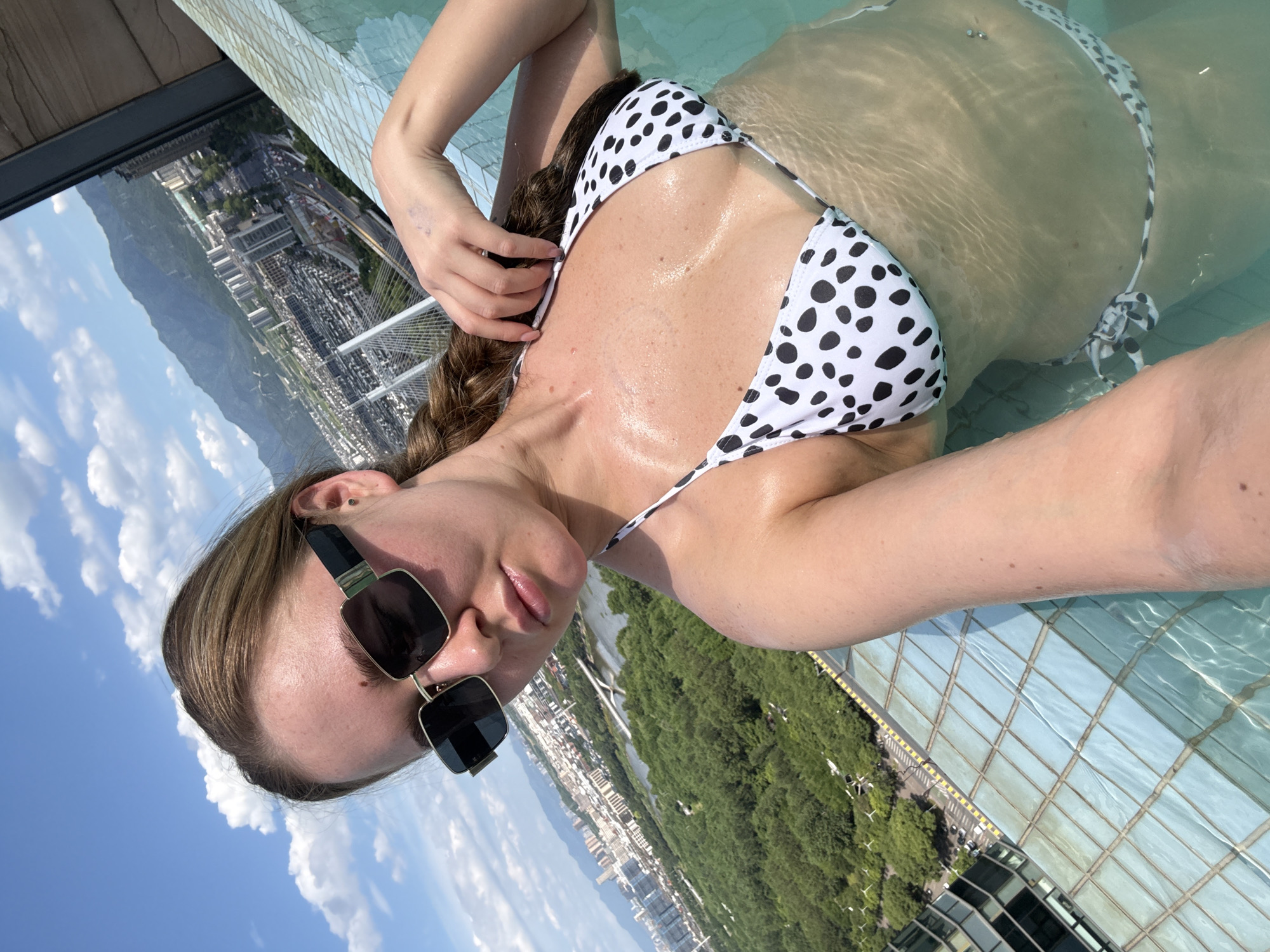 A woman is taking a selfie in an infinity pool overlooking the city of Yiwu, China. She has blonde hair in a braid and is wearing large, square, black-rimmed sunglasses and a white bikini with black polka dots. The pool is clear blue, and the water is rippling. In the background, there is a sprawling cityscape with buildings and lush green trees, and in the distance, mountains are visible. The sky is a clear blue with scattered white clouds. The woman is holding her hair with her right hand and her body is partially submerged in the water.