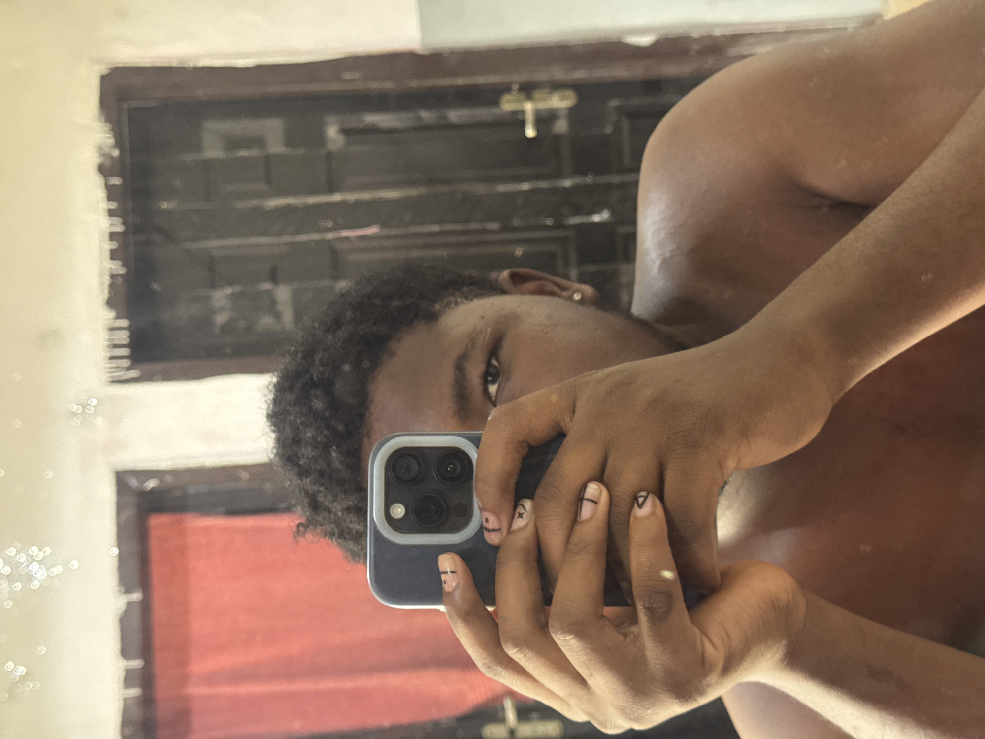 A person is taking a selfie in what appears to be a bathroom setting. The person has dark, curly hair and is holding a smartphone with multiple rear cameras. Their right hand is prominently displayed, revealing short fingernails with minimalist black line art. The person appears to be shirtless. The background consists of a mirror showing reflections of a room; the reflections include window bars and a section of red fabric. The setting is not distinctly recognizable as the city of Ipetumodu, Nigeria; location is not definitively identifiable.