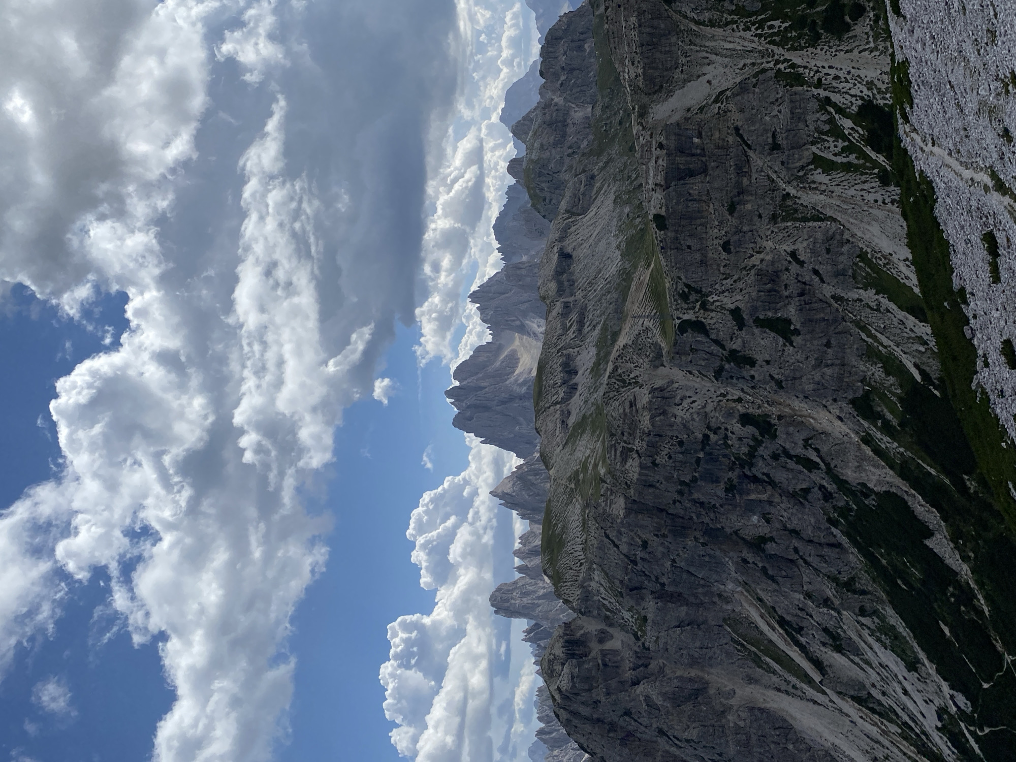 The image shows a panoramic view of a mountainous landscape in the Dolomites, likely near Auronzo di Cadore, Italy. The foreground is dominated by rocky slopes covered in sparse vegetation. In the mid-ground, a rugged mountain range with sharp peaks stretches across the frame. The background reveals more distant mountain formations fading into the haze. The sky is filled with dramatic, puffy clouds against a bright blue backdrop. The scene suggests a clear day with dynamic cloud formations, characteristic of high-altitude mountain environments. There are no people or man-made structures visible in the image, emphasizing the natural and remote character of the location.
