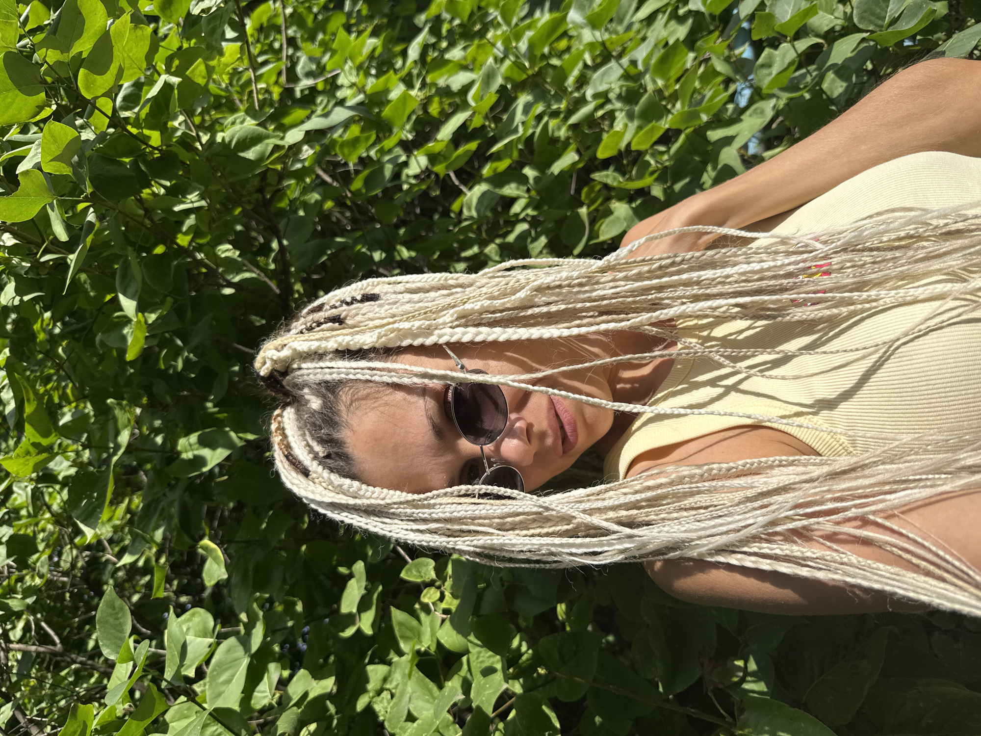 In Rusanovo, Russia, a woman with blonde box braids stands outdoors, partially obscured by lush green foliage. She is wearing a yellow ribbed tank top and dark oval sunglasses. The natural sunlight casts shadows across her face and arms, highlighting the texture of her braids. The background consists of dense green leaves and branches, suggesting a park or garden setting.