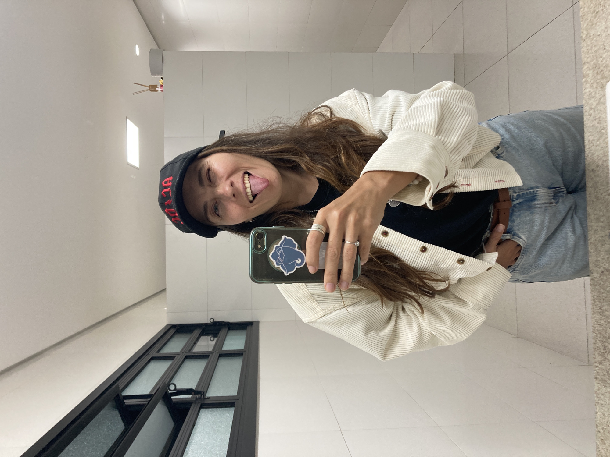 The image shows a woman taking a selfie in a bathroom. She is wearing a black baseball cap with red lettering, a black t-shirt, a white corduroy jacket, and blue jeans. Her tongue is sticking out, and she is winking. She is holding a phone with a dark green case. In the background, there is a window with a black frame, white tiled walls, and a white tiled floor. There is also a small window with an air freshener nearby. The location is likely in Florianópolis, Brazil, but this cannot be verified from the visual elements.