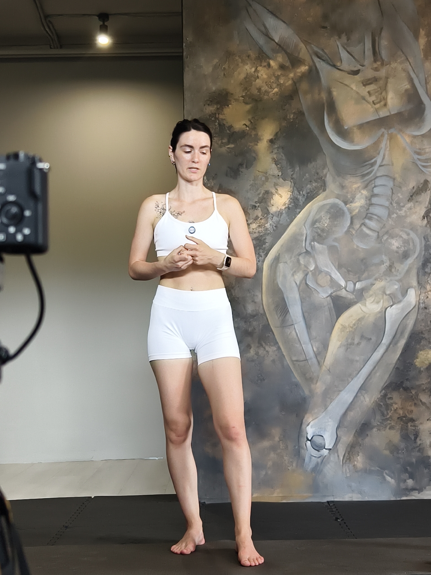 A young woman with dark hair pulled back stands in a room in front of a large mural that depicts skeletal figures. She is wearing a white sports bra and white shorts. Her hands are clasped in front of her. A camera on a tripod is positioned to her left, out of focus. The background consists of a plain wall and the mural. The floor appears to be a dark mat. The scene does not contain any specific indicators that would place it in Tyumen, Russia.