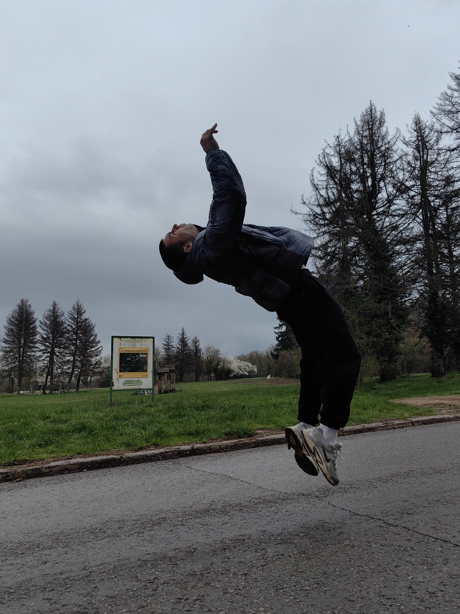 A man is in the air, performing a backflip on an asphalt road. The man is wearing black pants, a gray jacket, white socks, and sneakers. He is facing upward with his right arm extended. The background shows a grassy area with trees and a sign written in Bulgarian. The sky is overcast. The location is Shumen, Bulgaria, based on the Bulgarian text on the sign.