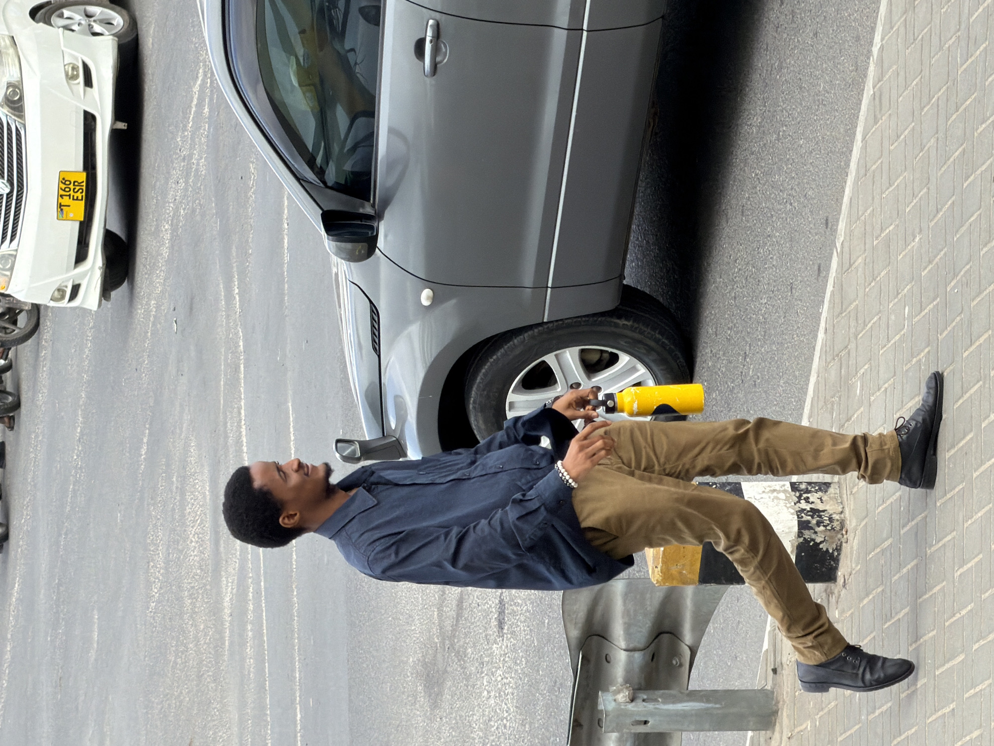 A man walks along a paved sidewalk next to a road in Dar es Salaam, Tanzania. He is wearing a dark blue collared shirt, brown pants, and black shoes. He carries a yellow water bottle in his right hand and wears a beaded bracelet on his left wrist. A gray SUV is parked to his right, and a white van with a yellow license plate is visible in the background. The sidewalk is made of interlocking pavers, and a metal guardrail runs along the edge of the road. The man is looking to his right with a slight smile.