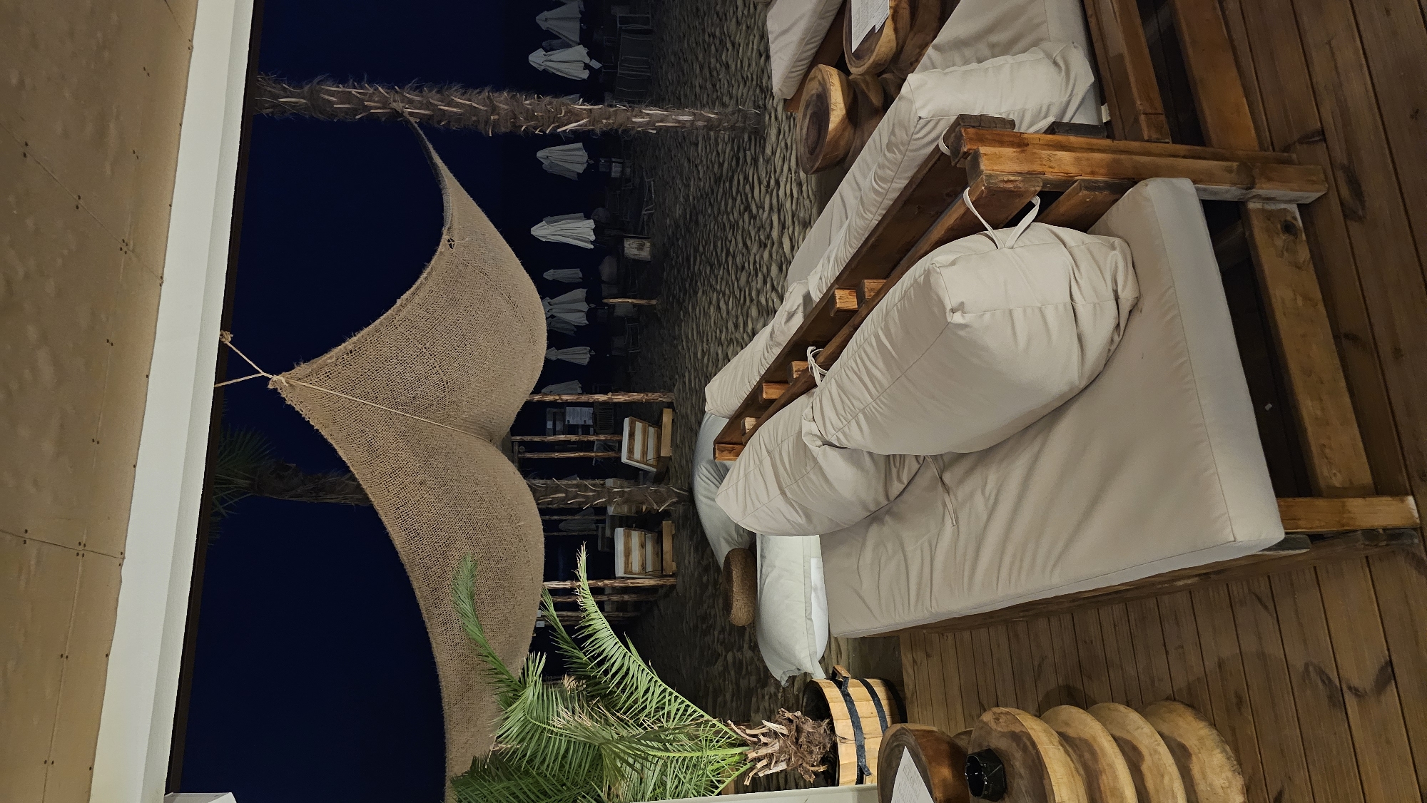The image depicts an outdoor lounge area on a sandy beach at night, likely in Varna, Bulgaria. In the foreground, there are several low wooden lounge beds with light-colored cushions and large pillows. To the left, a small potted palm tree stands on a wooden deck. In the background, rows of closed white beach umbrellas are visible on the sand, with more lounge furniture and palm trees in the distance. A thatched-style canopy provides shade over the lounge area. The lighting suggests it is dusk or nighttime. No people are present.