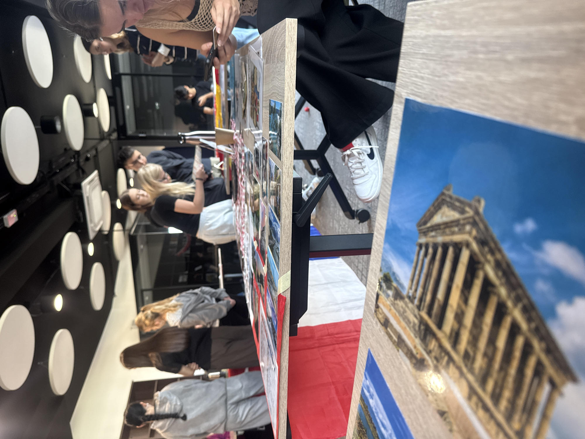 The image depicts a group of young adults gathered around tables displaying various photographs and informational materials. The environment appears to be an indoor space with a modern aesthetic, characterized by black ceilings with circular acoustic panels and track lighting. Several individuals are seen interacting with the displays, some looking down at the materials while others engage in conversation.
One table in the foreground is covered with several large, glossy photographs, one of which shows a close-up view of ancient stone structures against a bright blue sky. Another photograph appears to depict a landscape with water. A portion of a French flag is visible draped over the edge of this table.
In the middle ground, another table is laden with numerous smaller photographs and documents, laid out in a grid-like pattern. Several people are leaning over this table, examining the content. A young woman in a black top and white sneakers is seated in the foreground, looking at her phone. Behind her, other individuals are visible, including a young woman with blonde hair and a black t-shirt, and a young man in a dark shirt.
The background reveals more of the indoor space, with shelves and glass partitions suggesting it might be an office, a classroom, or a communal area. The presence of the French flag suggests a connection to France, possibly an event or exhibition related to French culture or travel. Without further information, it is not possible to determine the specific location in Opole, Poland.
