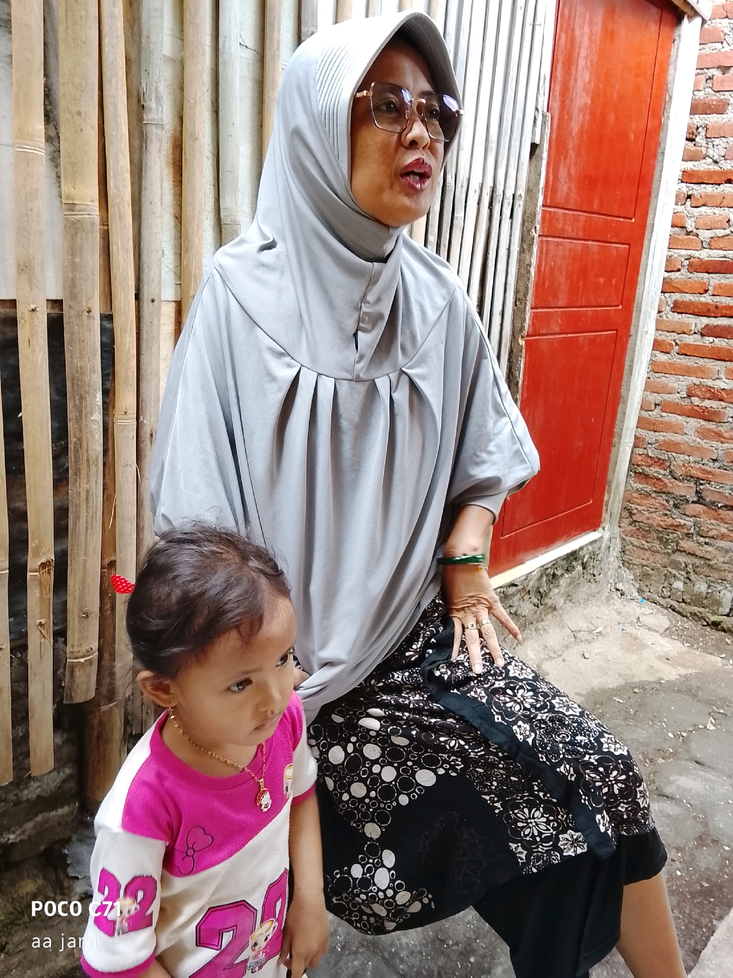 A medium shot shows a woman wearing a grey hijab and glasses, seated with a young child in front of her. The woman is looking to her right, with her mouth slightly open. She is wearing a long grey garment over a black and white patterned skirt. She has a bracelet on her right wrist and rings on her fingers. The child, wearing a pink and white shirt, is looking to their left. The background consists of a bamboo wall on the left, a red door in the center, and a brick wall on the right. The scene appears to be an outdoor setting. The text "POCO C71 aa jami" is overlaid on the bottom left of the image.