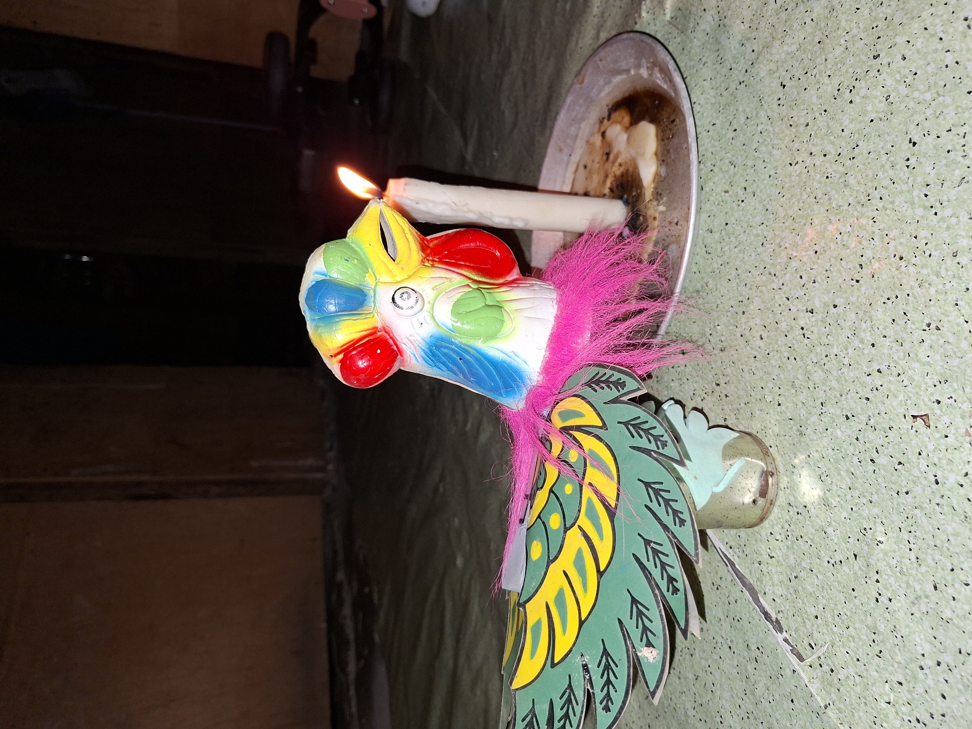 A brightly colored, toy bird-shaped candle holder is positioned on a green surface next to a lit candle. The holder is predominantly green, yellow, red, white, and blue. A pink, feathery fringe adorns the top of the bird. The candle is set in a shallow, round metal dish. The background is blurred, displaying a dark interior room. This location is in Lhokseumawe, Indonesia.