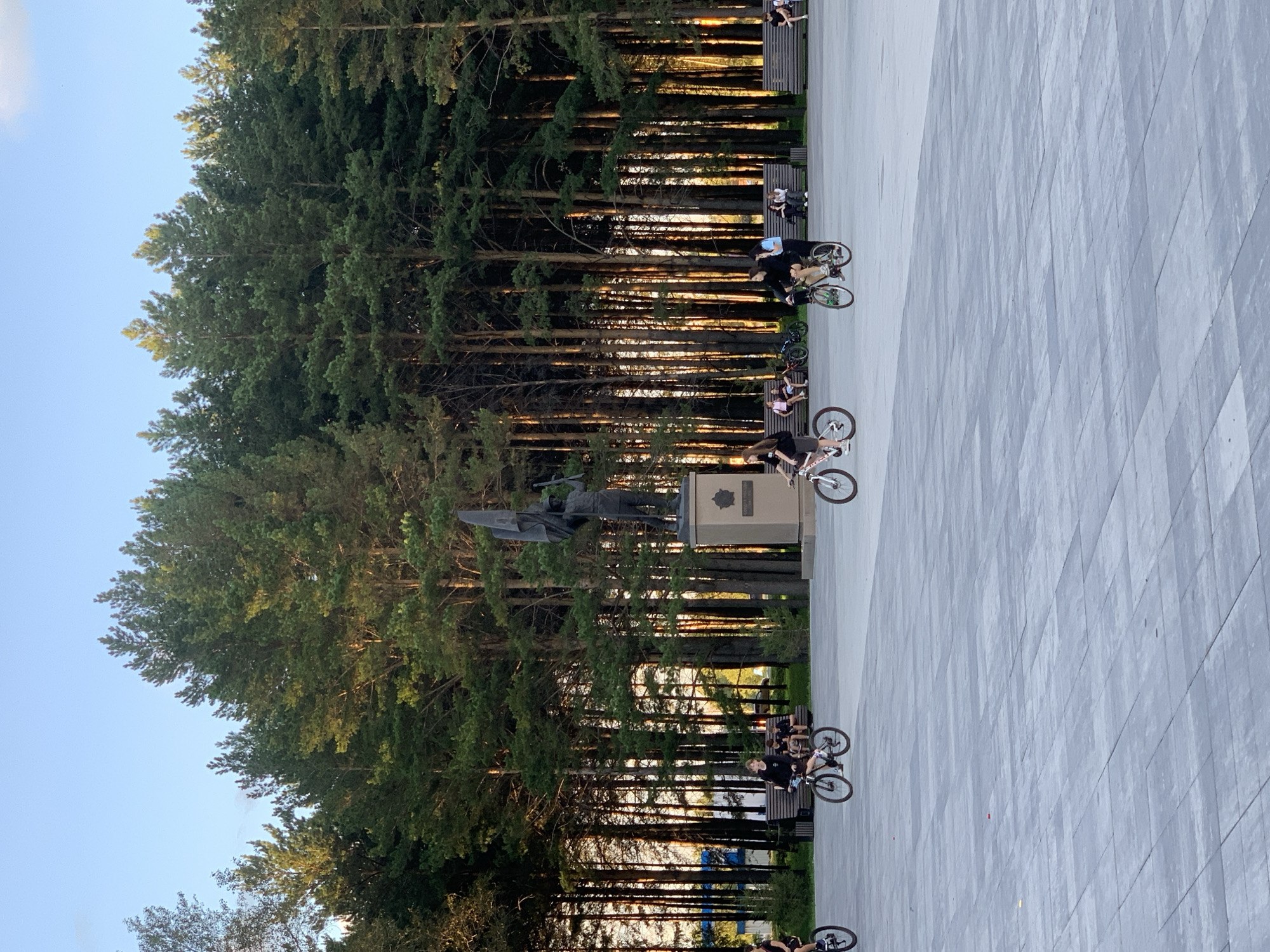 A wide paved plaza is in the foreground, with a dense forest of tall pine trees forming the background. In the middle ground, several young people are riding bicycles. To the right of the plaza, a bronze statue of a soldier holding a flag stands on a pedestal. A park bench is visible to the left of the statue, and other benches are further back near the trees, occupied by seated individuals. The scene appears to be in a park or public square in Prokopyevsk, Russia, likely during daylight with the sun casting warm light through the trees.
