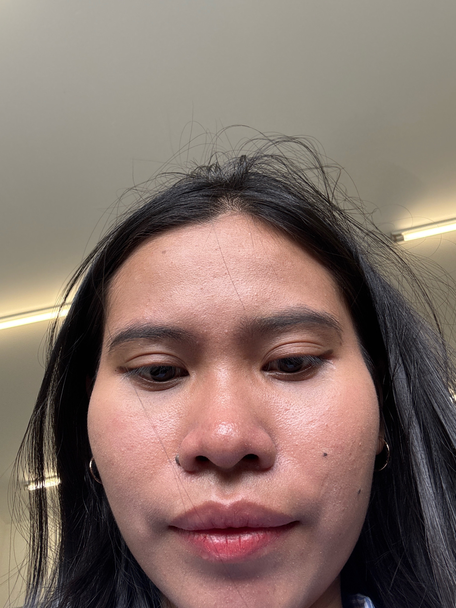 The image shows a close-up, low-angle shot of a young woman's face, with her forehead and the top of her head filling the upper portion of the frame. She has dark, long hair that is slightly disheveled. Her skin appears to have some texture and small blemishes. She is wearing small hoop earrings. The background consists of a light-colored ceiling with fluorescent lighting fixtures visible. The scene is likely indoors, possibly in a public space or a home, given the ceiling and lighting. There is no indication of the specific location within East Jakarta, Indonesia, beyond the general context provided.