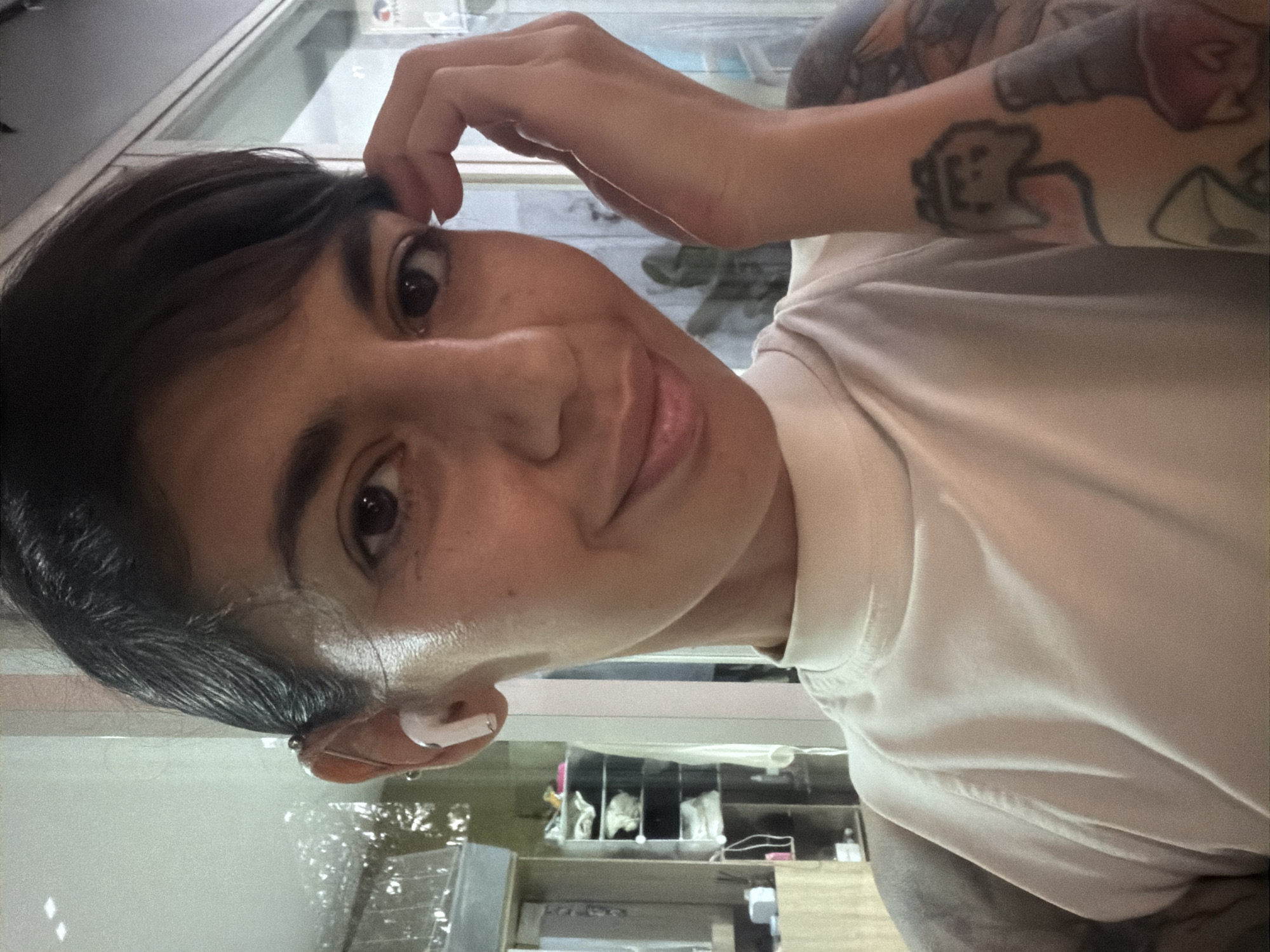 A person with dark hair, styled with bangs, and a medium skin tone is depicted in a close-up, selfie-style photograph taken indoors in Kuta Selatan, Indonesia. The individual, appearing to be female, looks directly at the camera with a gentle smile. They are wearing a light-colored, high-neck, sleeveless top. A white wireless earbud is present in the left ear, which also features a silver industrial bar piercing in the upper cartilage. The person's left hand is raised to the side of their head, fingers lightly touching the temple area. The right arm, partially visible on the left side of the frame, is heavily tattooed with colorful, cartoon-style imagery, including a distinct pixelated video game controller design in gray and pink, and other abstract shapes in red and orange. The background is softly blurred but reveals an indoor setting with shelves containing various items, reflections in a glass surface, and ambient artificial lighting.