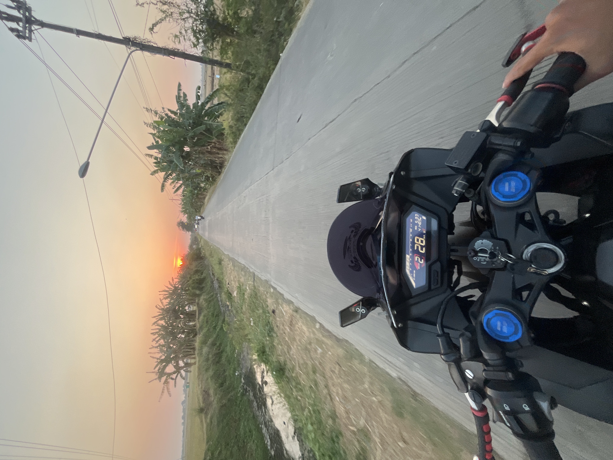 The image captures a first-person perspective of someone riding a motorcycle on a paved road at sunset. The road is lined with vegetation, including banana trees, and a ditch with water on the left side. In the distance, the sun is setting on the horizon, casting a warm glow over the scene. There are utility poles with wires along the side of the road, and a few other motorcycles are visible further ahead. The motorcycle's dashboard shows a speed of 28 km/h and a time of 5:33. The location is identified as Batukali, Indonesia.