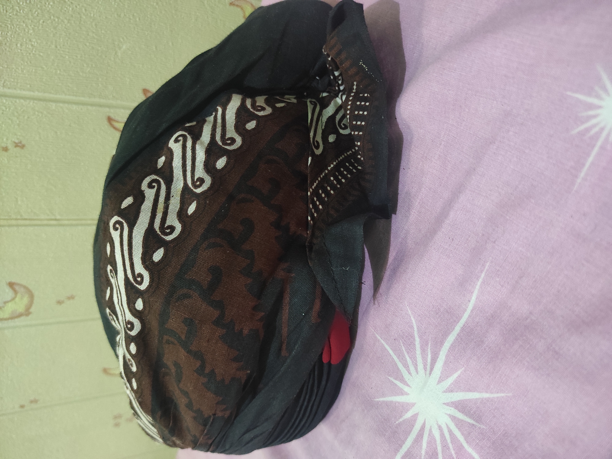 A batik-patterned head covering, possibly a traditional Javanese "blangkon," rests on a pink, star-patterned cloth. The head covering features a brown base with intricate white and black batik designs, some resembling swirls and floral motifs. A portion of red fabric is visible underneath the batik material on the left side. The background is a textured, light-colored wall with faint, abstract yellow and light brown patterns. The scene appears to be indoors, possibly in a residential setting in Pleret, Indonesia. There are no people or actions depicted.