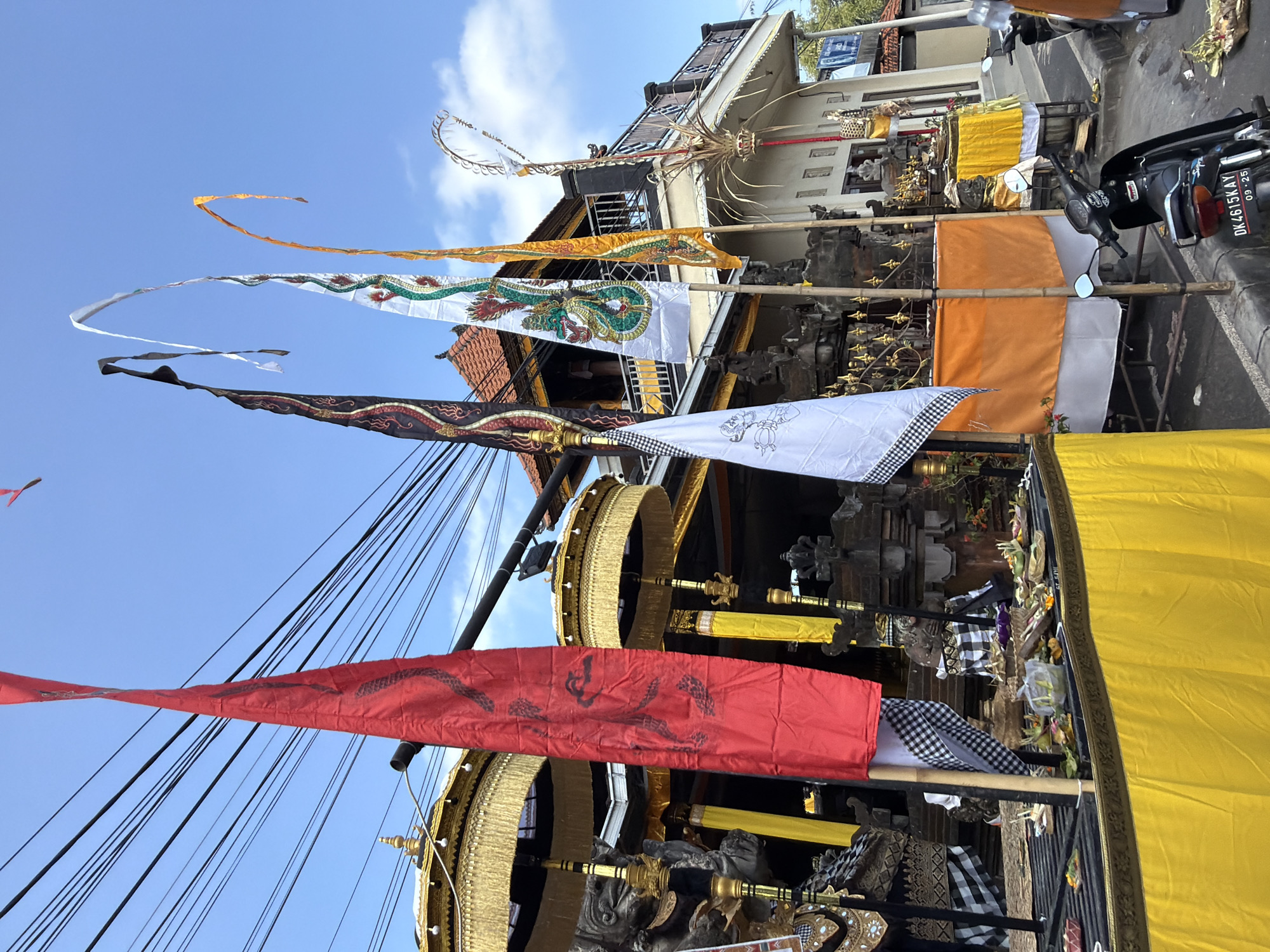In Blahbatuh, Indonesia, a scene unfolds with multiple flags, umbrellas, and offerings, likely for a religious or cultural ceremony. A large red flag with a black serpentine design hangs prominently, supported by a black pole. Behind it, two tiered, golden umbrellas with fringed edges are visible, suggesting a ceremonial context. Several other decorative flags are present: one black with intricate red and gold patterns, a white flag adorned with a green and red dragon, and a yellow and orange flag with a similar dragon motif. A white flag with a black and white checkered pattern is also visible.
The background reveals a building with traditional architecture, featuring red-tiled roofs and ornate detailing. Several small stone statues or carvings are incorporated into the structure and surrounding areas. A table or platform is laden with offerings, including food items, flowers, and woven baskets, arranged on green leaves. The ground in front of the offerings is covered with a bright yellow fabric.
In the lower right portion of the image, a black motorcycle with a license plate reading "DK4615KAY 09-25" is parked. Further to the right, part of another motorcycle or scooter is visible. The environment appears to be a street or pathway, with overhead power lines stretching across the blue sky. The overall scene is a vibrant display of cultural elements, suggesting a significant event is taking place or has recently occurred.