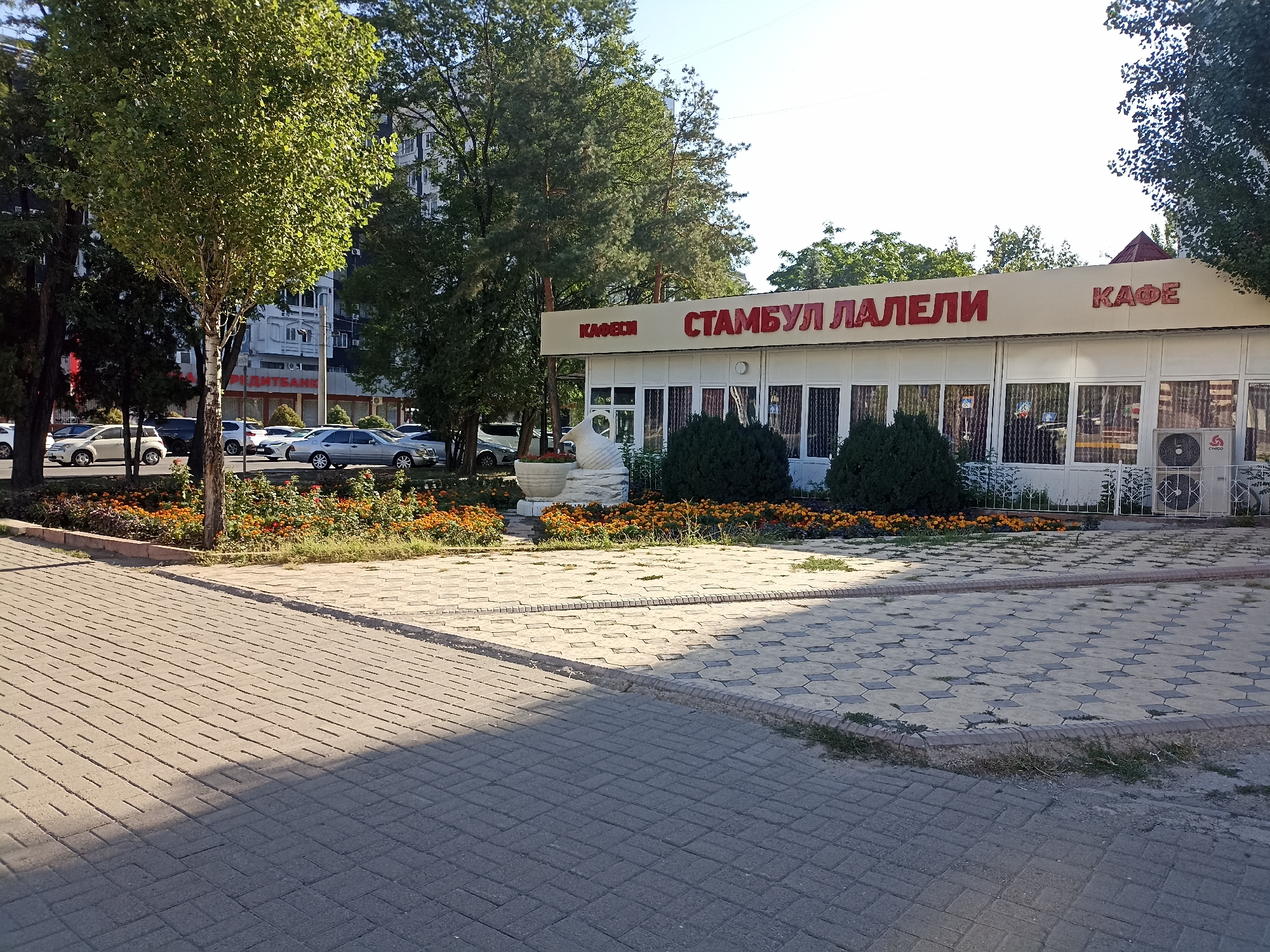 The image depicts the exterior of a cafe named "Стамбул Лалели" (Istanbul Tulip) in Bishkek, Kyrgyzstan. The cafe has a light-colored facade with red lettering and a row of windows. In front of the cafe, there are some bushes and a decorative sculpture. The ground is paved with interlocking concrete blocks. Several cars are parked on the street in the background. Lush green trees partially obscure the view of buildings behind the cafe. The sky is clear and blue, indicating it is daytime.
