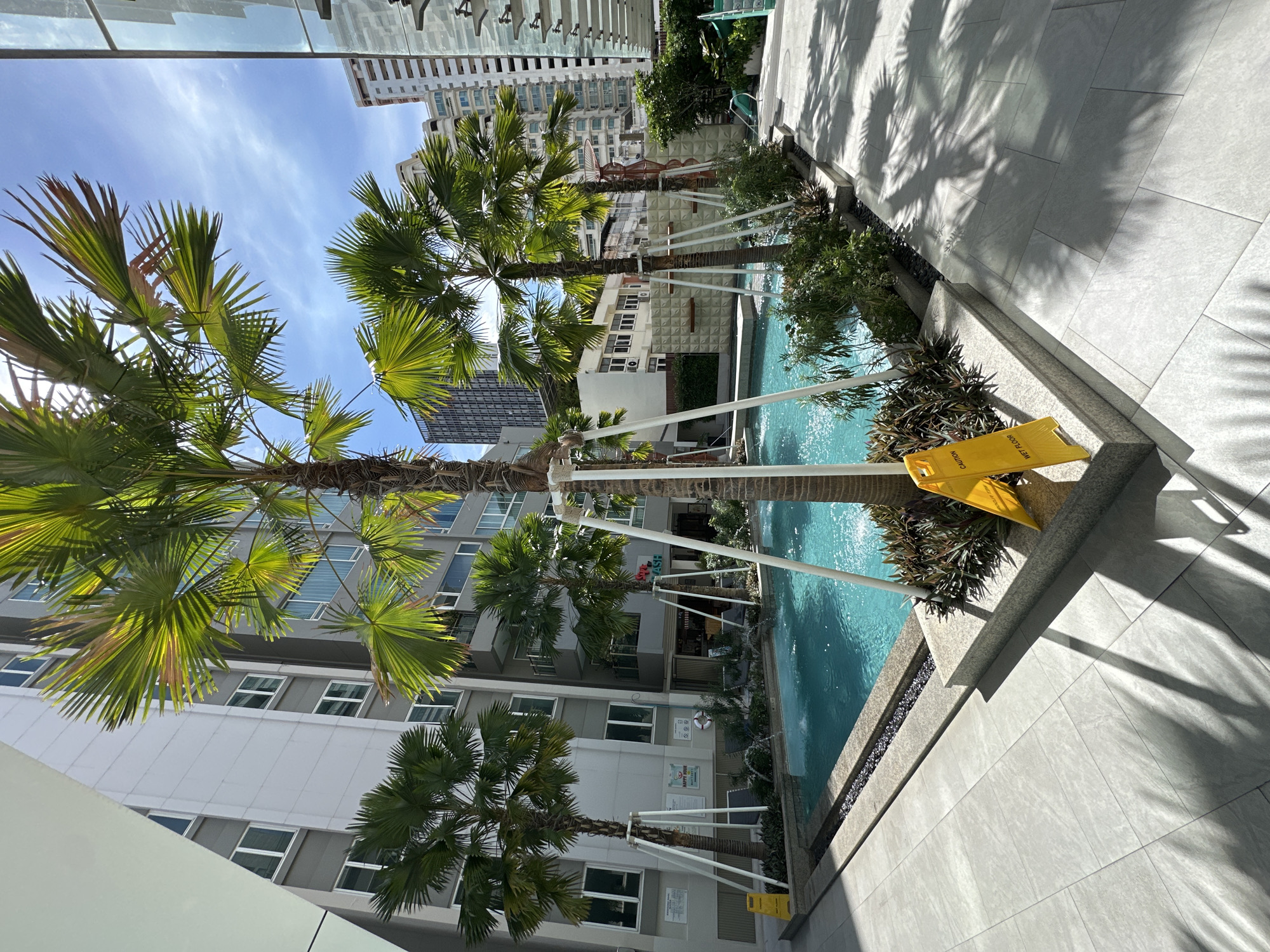 This outdoor daytime shot, taken at a high angle, depicts a swimming pool area in Bangkok, Thailand. The pool is surrounded by palm trees and is situated between tall buildings under a partly cloudy sky. A yellow "CAUTION WET FLOOR" sign is visible near the edge of the pool. Shadows from the palm trees are cast on the light-colored tiled ground. The scene suggests an urban recreational space within a densely built environment.
