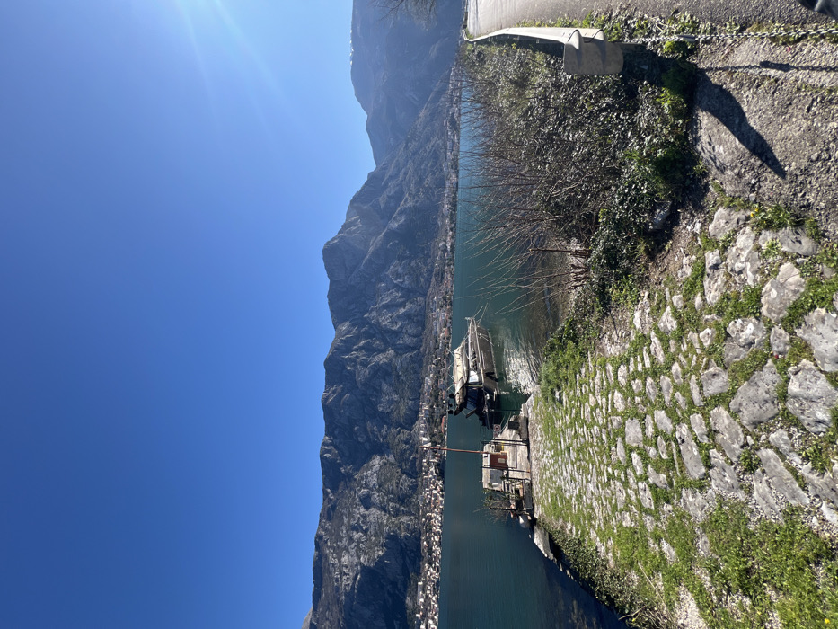 The image depicts a sunlit coastal scene captured in Bogdašići, Montenegro, featuring a body of water bordered by a mountainous landscape. In the left foreground, a stone-paved path, partially covered with green grass, descends toward the water. This path terminates at a small, rectangular concrete pier where a dark-hulled motor yacht, equipped with a light-colored canopy, is moored. To the right of the paved path, a strip of sparse vegetation and bare ground runs parallel to the water, adjacent to a paved road. This road is delineated by a silver metal guardrail on its left edge. Across the calm, greenish-blue water, a densely settled town with multiple buildings is visible along the distant shoreline. Behind the town, a prominent range of rugged, grey mountains rises steeply, dominating the background. The sky above is uniformly clear and bright blue. Direct sunlight from the upper right generates a visible lens flare and casts distinct shadows on the foreground elements. No individuals are present in the scene.