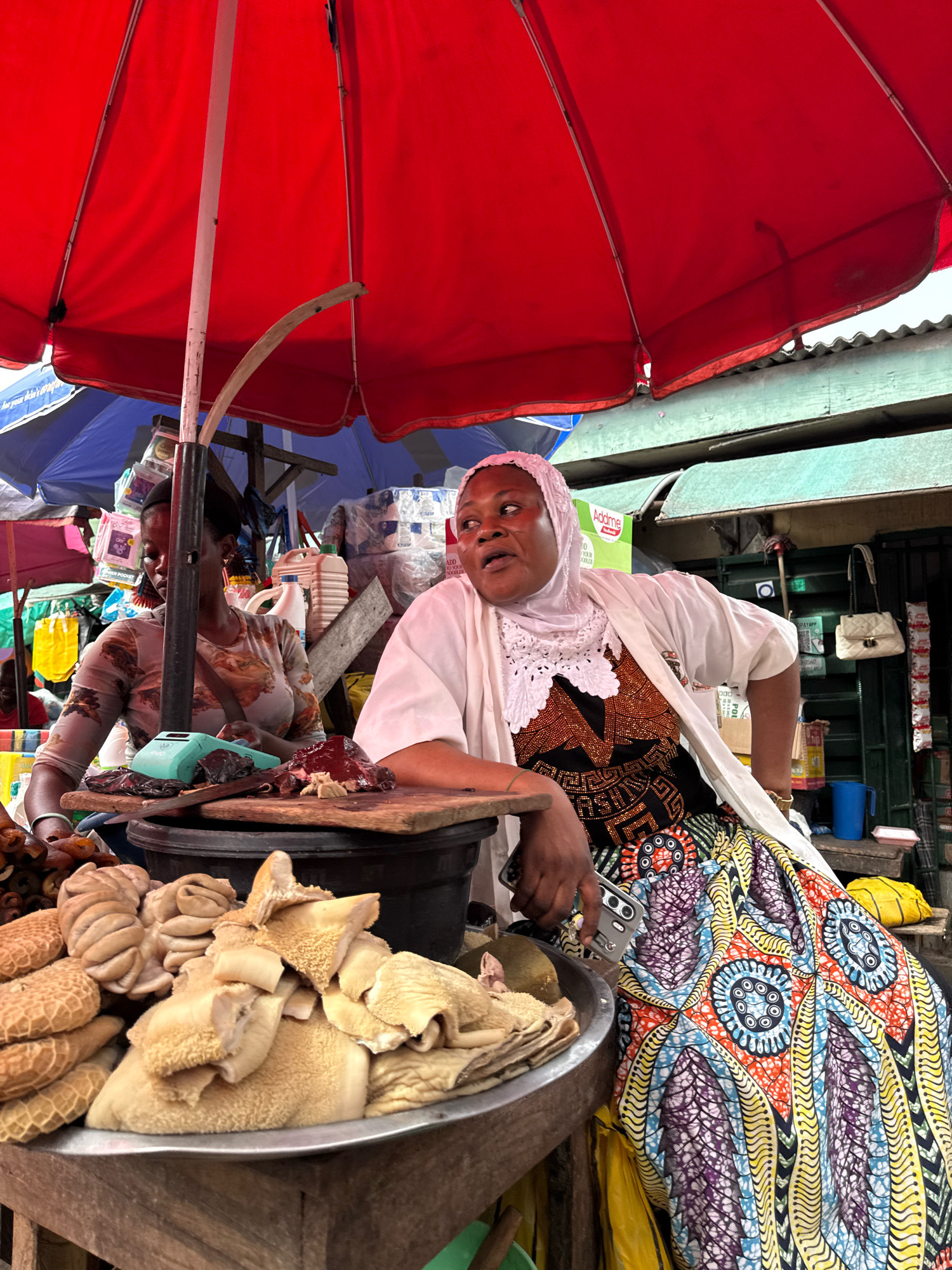 A market scene in Lagos, Nigeria, is depicted under a large red umbrella. Two women are present at a stall. The woman on the right, wearing a light-colored headscarf and a white open-fronted robe over a black and orange patterned garment and a colorful, vibrant patterned skirt, is looking off to her left and speaking. The woman on the left, wearing a patterned top, is looking down at a teal-colored scale on a wooden surface. In front of the women, on a wooden board, are piles of meat, appearing to be tripe and other offal, displayed in a metal basin. Behind the women, the market environment is visible, with stacked goods, bags, and other stalls. The lighting suggests it is daytime.