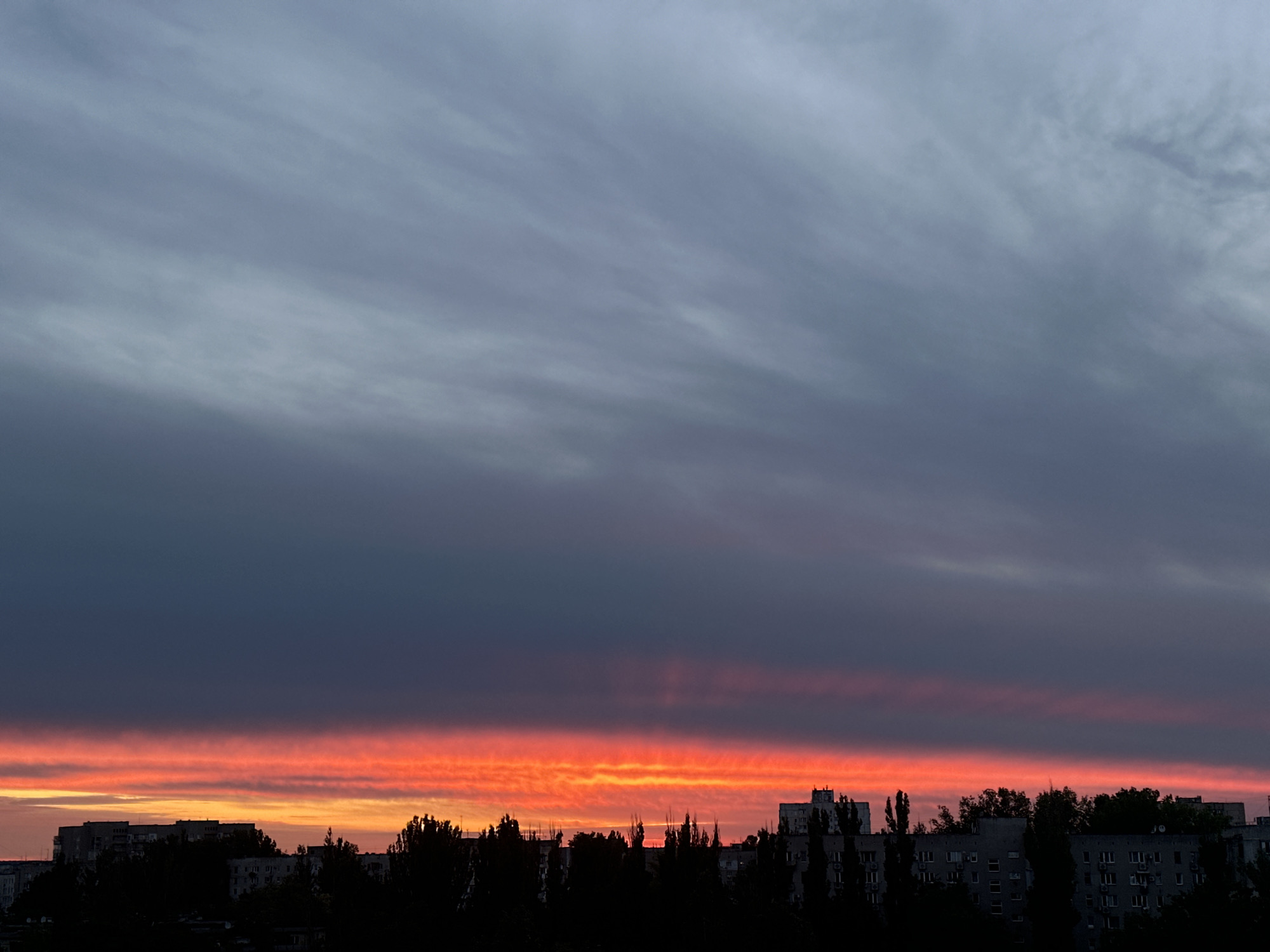 The image depicts a cityscape at dusk or dawn in an unidentified location. The sky is dominated by layers of clouds, transitioning from a pale, overcast grey at the top to a vivid band of red and orange hues along the horizon. The lower portion of the image is filled with the silhouetted outlines of buildings and trees, creating a stark contrast against the colorful sky. The buildings are a mix of low-rise structures and taller apartment blocks, typical of urban residential areas. The trees are interspersed among the buildings, adding to the urban landscape. No people are visible in the scene. The image captures a moment of natural beauty in an otherwise ordinary urban setting.