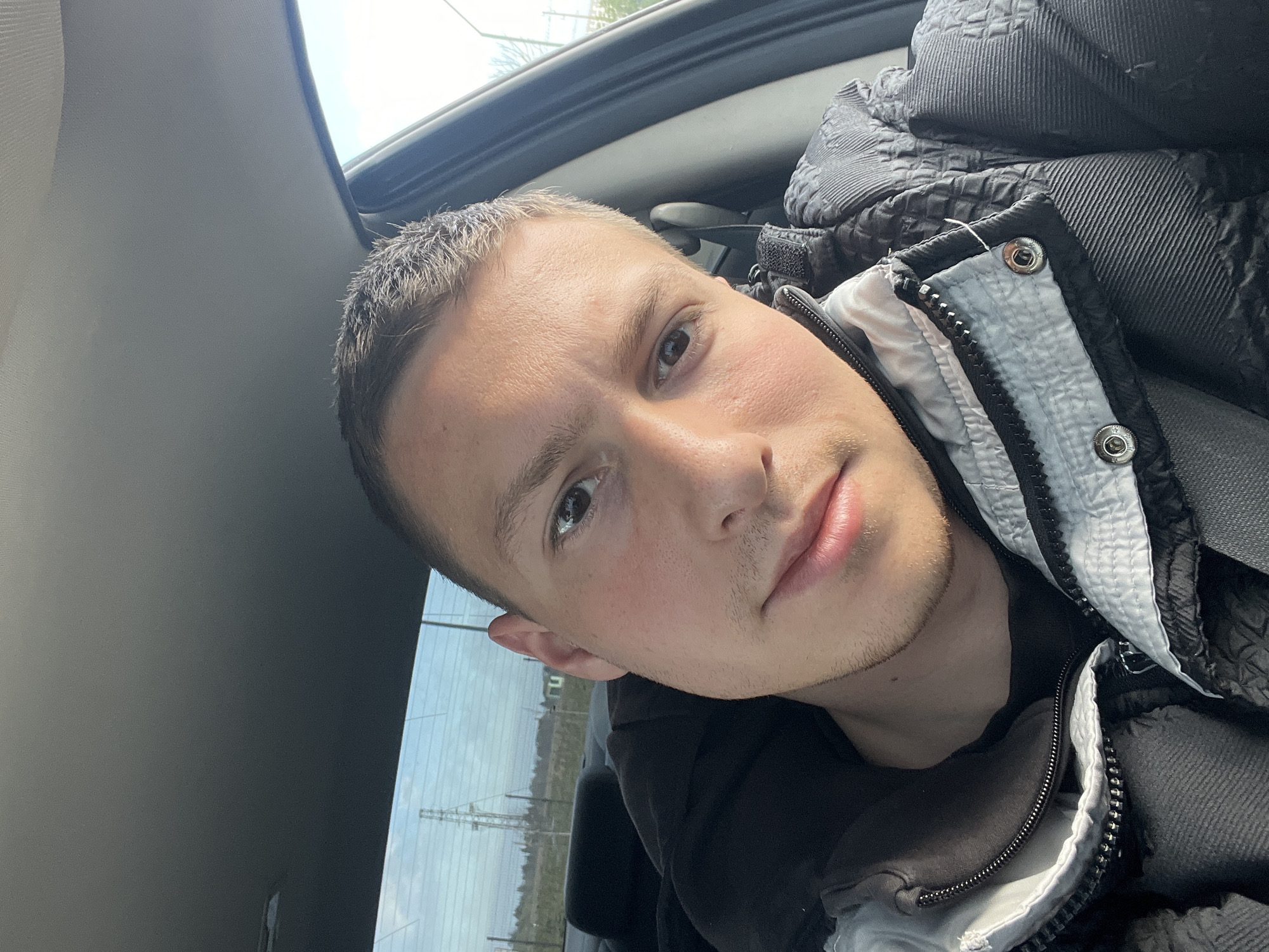 The image is a close-up selfie of a young man in what appears to be a car. He has short, light brown hair and is wearing a dark jacket with a light-colored lining. The jacket's zipper is partially open. The interior of the car is visible, including the roof and window. Outside the window, there are power lines and a glimpse of buildings and greenery. The location is potentially in Burgas, Bulgaria, though specific landmarks are not visible for positive confirmation from this image alone. The man's gaze is directed at the camera. The environment appears to be daytime with diffused sunlight. No interactions or actions can be determined beyond the man taking a selfie.
