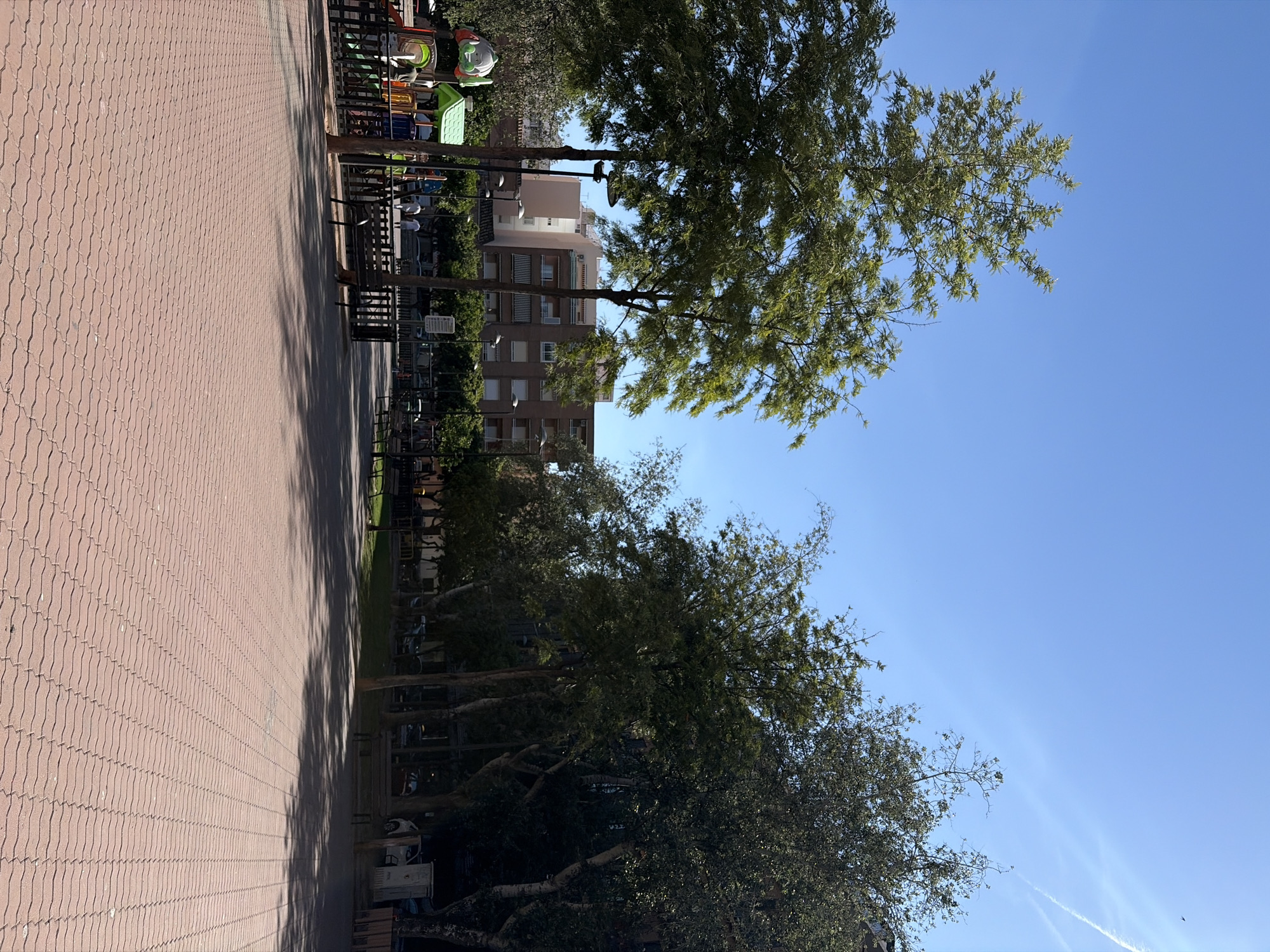 The image shows a park in Sagunto, Spain, featuring a red brick pavement on the left side. Trees line the right side, casting shadows on the pavement. A building is visible in the background, partially obscured by the trees. A playground with colorful equipment is located near the trees. The sky is clear blue with some airplane trails. There are benches and fences along the trees.