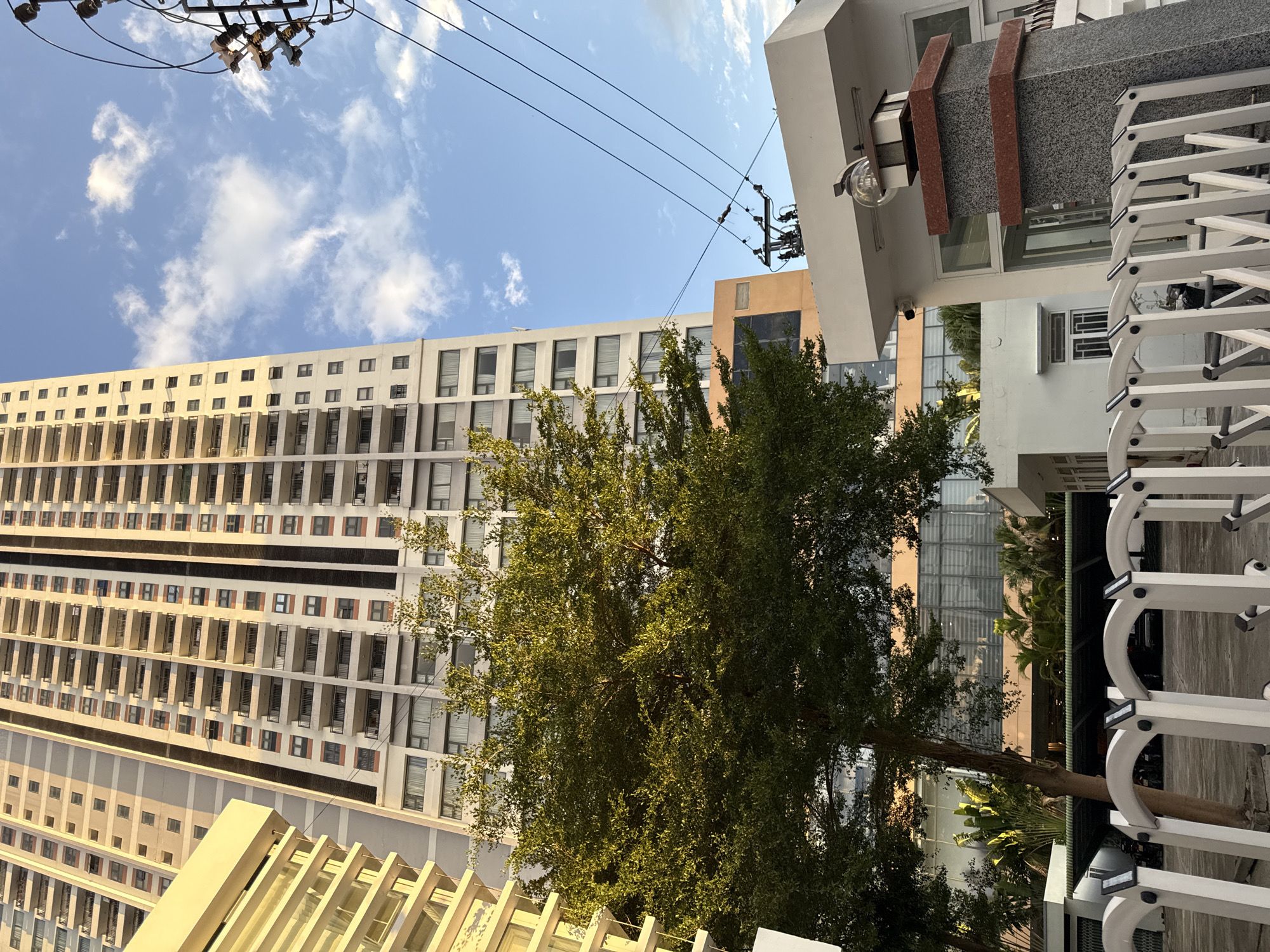 The image shows a tall, multi-story building in the city of Nha Trang, Vietnam. The building is light-colored with many windows and balconies. In the foreground, there is a white metal gate and a large green tree. A utility pole with multiple wires is visible on the right side of the image. The sky is blue with some white clouds. The overall scene depicts an urban environment with a mix of modern architecture and greenery.