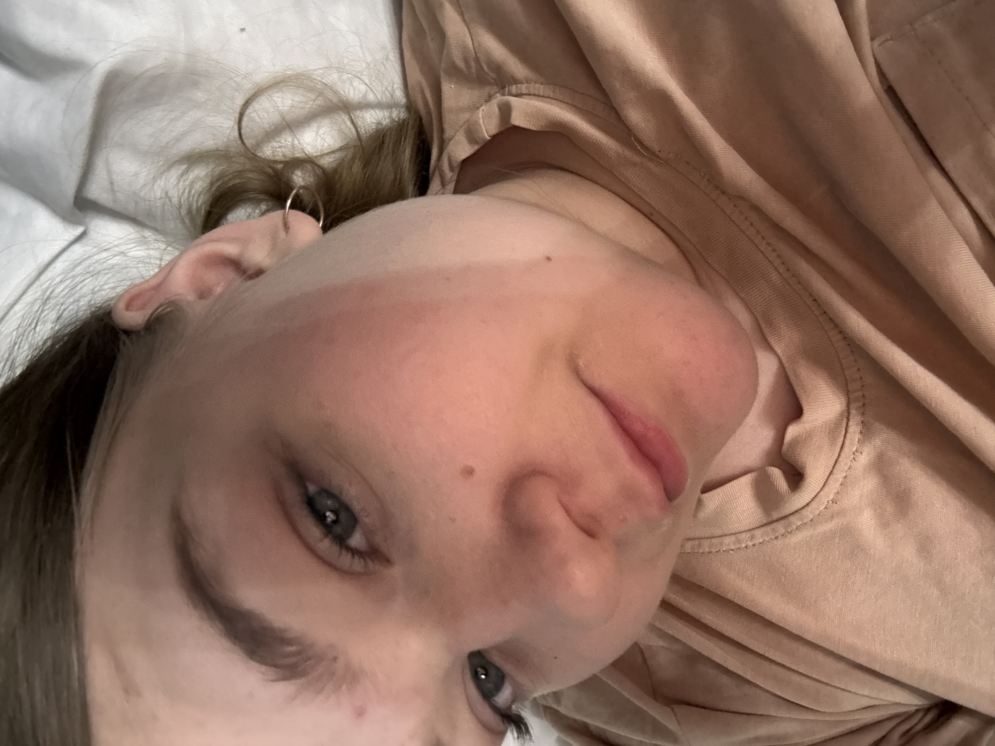 A close-up photograph depicts a young woman with fair skin, light brown hair, and blue eyes. She is wearing a light brown shirt. A small hoop earring is visible in her left ear. The background appears to be white bed sheets. The location is not discernible from the image. No information about the city of Chaikovsky, Russia can be inferred from the provided image.