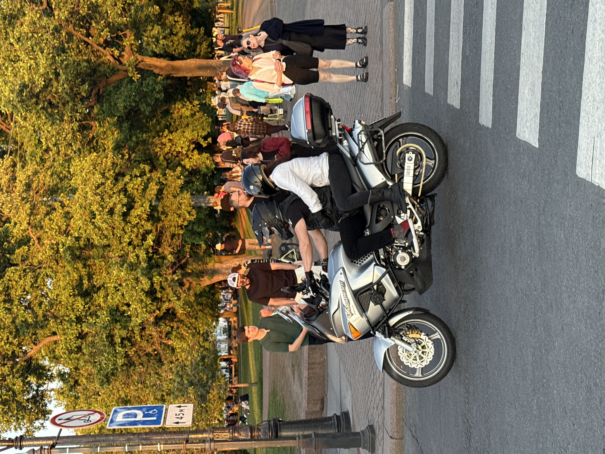 This image captures an urban street scene in Saint Petersburg, Russia, during the golden hour, evidenced by the warm, soft light illuminating the trees and subjects.
In the center foreground, a silver-grey Honda Varadero touring motorcycle, with "VARADERO" visible on its fairing, is positioned on an asphalt road. Two individuals are seated on the motorcycle, both wearing black full-face helmets. The driver is dressed in a black jacket, black pants, and red sneakers. The passenger, wearing a white long-sleeved shirt, black pants, and black boots, sits behind the driver. The motorcycle features a large windscreen, disc brakes, and a black top case at the rear.
To the left of the motorcycle, a black ornate streetlamp post stands, displaying three vertical signs: a circular white sign with a red diagonal bar over a scooter icon (indicating prohibition), a blue square sign with a white 'P' and a wheelchair symbol (indicating handicapped parking), and a white rectangular sign below it with "↑ 45м ↑" (indicating parking 45 meters ahead). Standing near the motorcycle on the left side are two men; one in a dark green polo shirt and dark pants, and another with a beard, wearing a white baseball cap, black t-shirt, and white shorts.
On the paved sidewalk to the right of the motorcycle, two women are standing. One woman, with prominent purple/pink hair, wears a light-colored collared shirt, black shorts, and black sandals. Beside her, an older woman with grey hair is dressed in a black long-sleeved outfit, black fishnet tights, and black flat shoes. Both women appear to be observing the motorcycle or its occupants.
The road surface in the lower right features white pedestrian crosswalk stripes. The background is dominated by a lush green public park, filled with numerous mature deciduous trees, some brightly lit by the setting sun. Many people are scattered throughout the park, walking, standing, or sitting, indicating a public gathering or leisure activity.