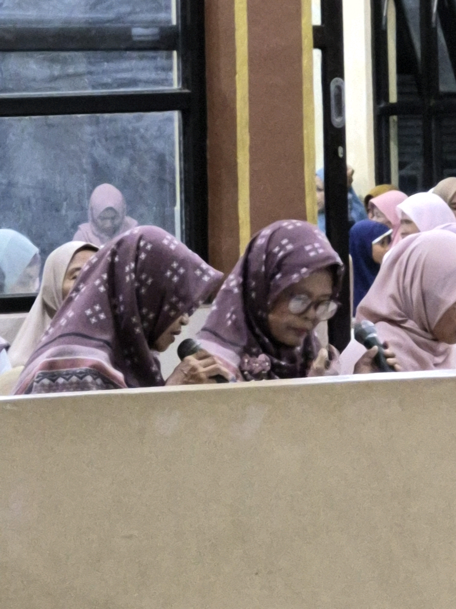 The image shows a group of women wearing hijabs. Two women in the foreground are speaking into microphones. The woman on the left is wearing a purple hijab with a white diamond pattern and a pink scarf. The woman on the right is wearing a purple hijab with a white diamond pattern and glasses. Behind them, other women are visible, also wearing hijabs in various colors including pink and light blue. They appear to be in an indoor setting with a brown wall and a window in the background. The context suggests a public gathering or event in Dukuhrejo, Indonesia.
