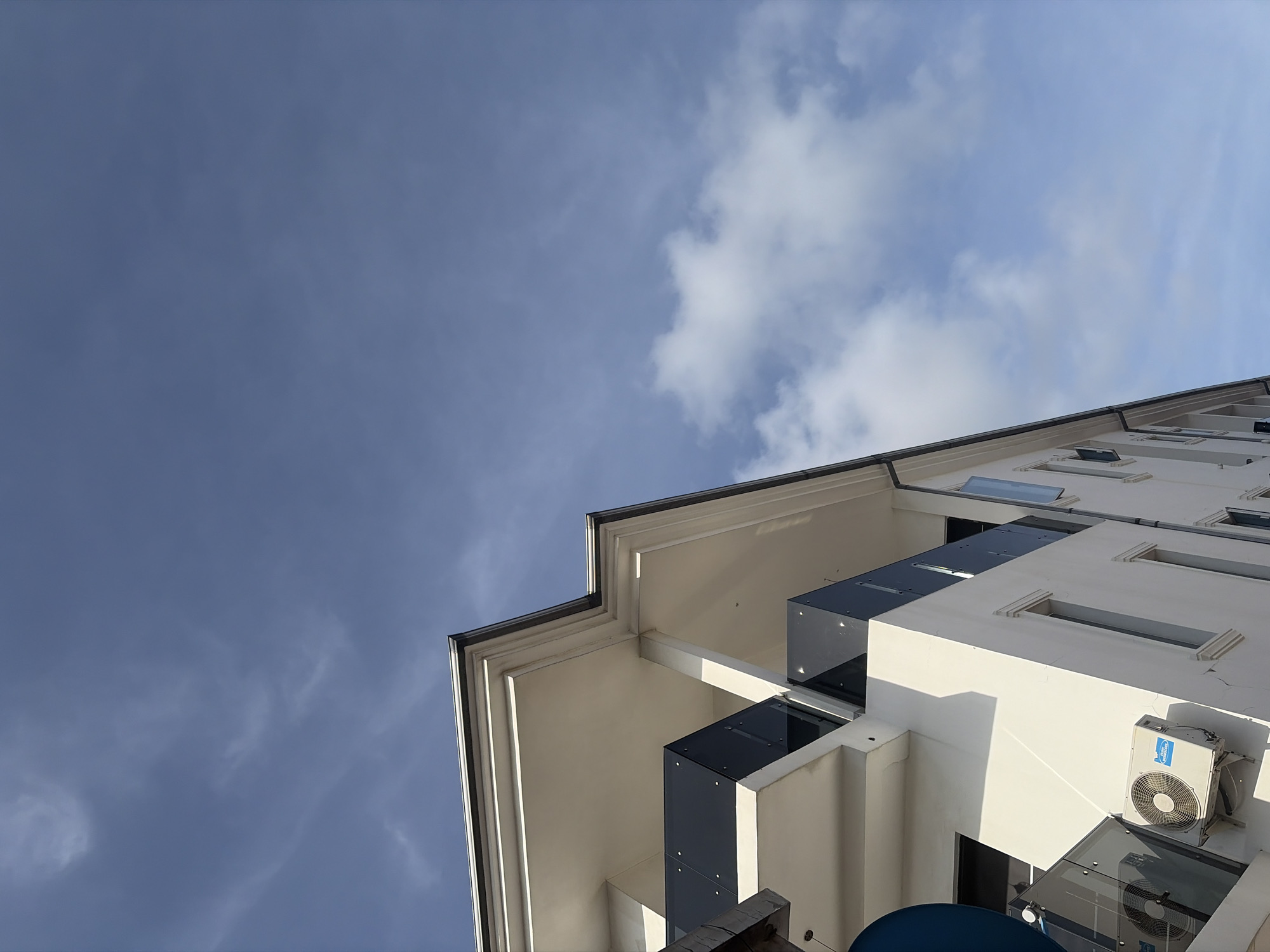 The image shows the exterior of a modern building in Igboefon, Nigeria. The building is white with dark glass balconies. An air conditioning unit is mounted on one of the lower walls. The sky is visible above, with a mix of blue and white clouds.